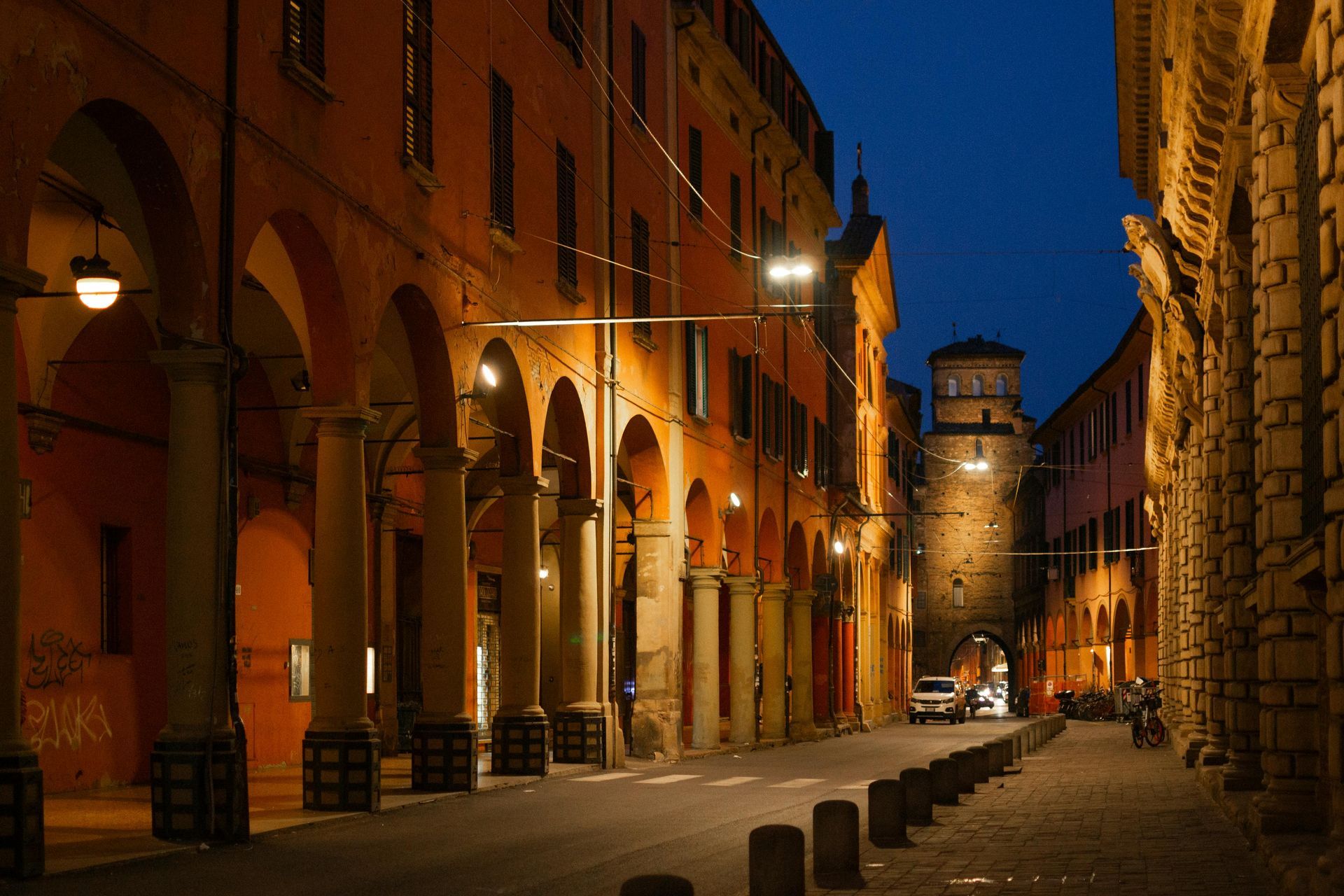 Cobblestone street lined with orange buildings and arches in Bologna, Italy at dusk. A few vehicles.