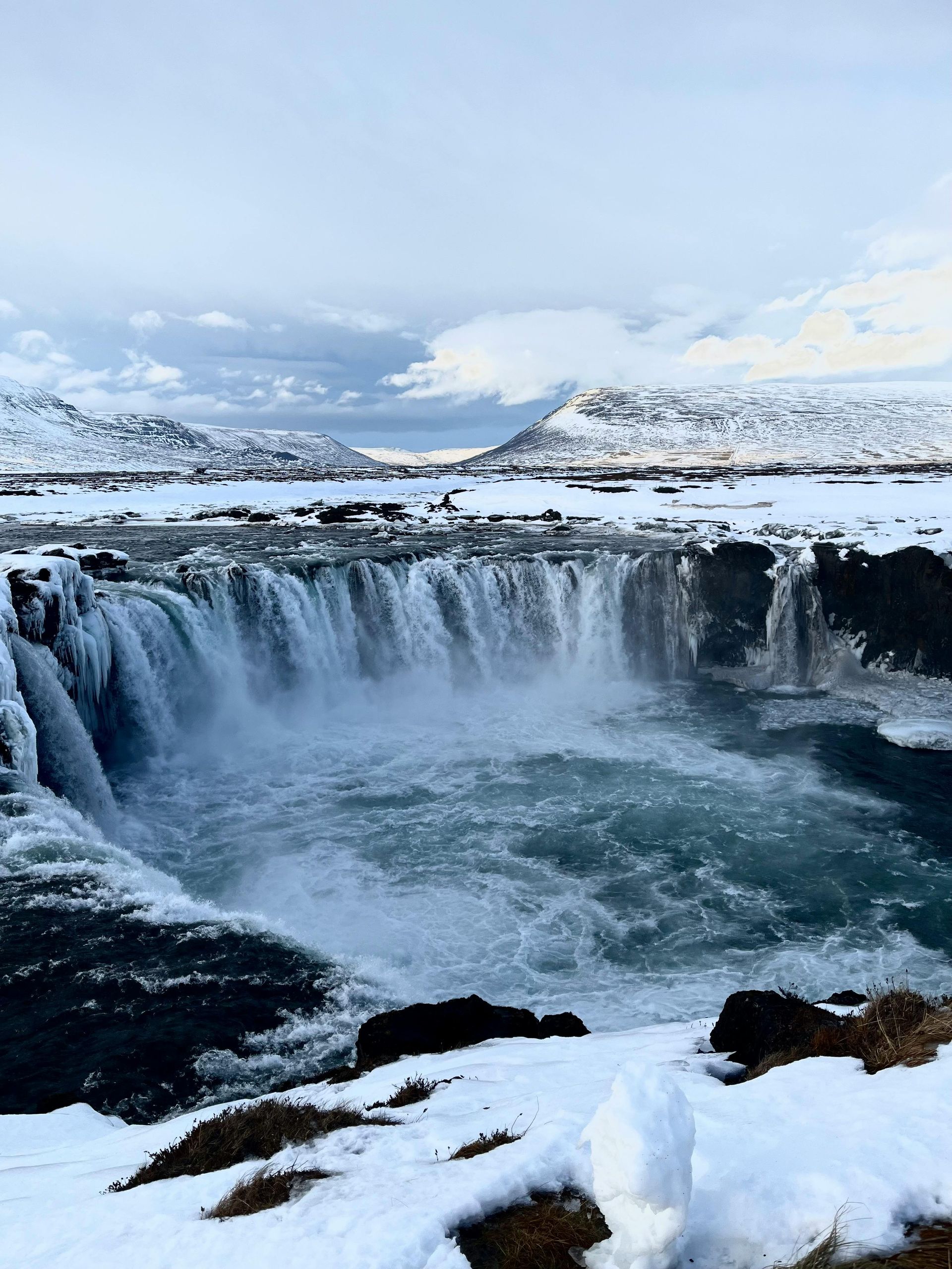 Waterfall cascading into a glacial pool, surrounded by snow-covered banks and mountains under a cloudy sky. Iceland