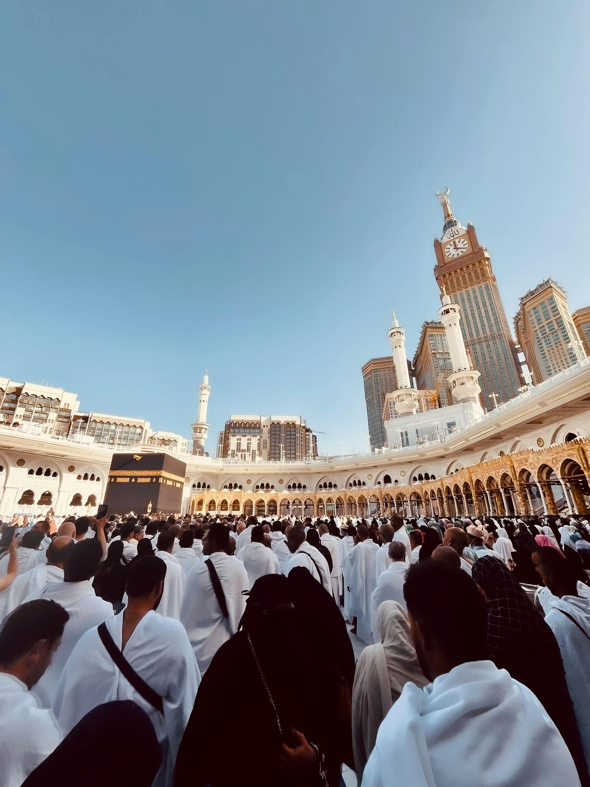 People in white robes circumambulating the Kaaba in Mecca, Saudi Arabia, with the iconic clock tower visible.