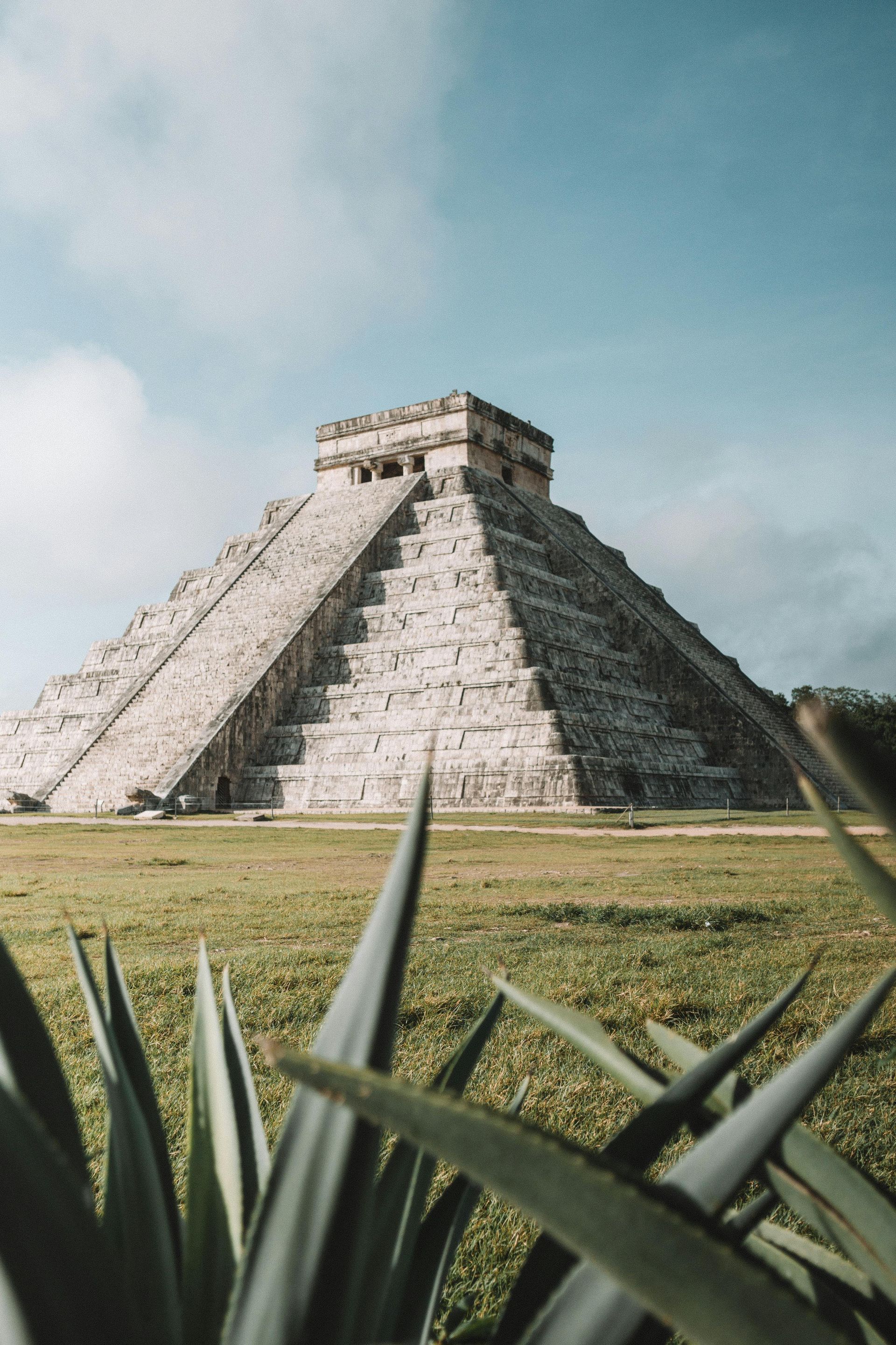 El Castillo pyramid in Chichen Itza, Mexico, seen from a grassy field, under a partly cloudy sky.