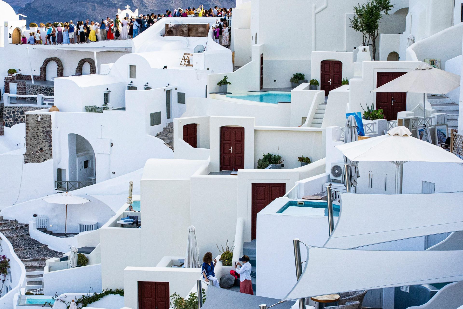 Whitewashed buildings with red doors, blue pools, and people on rooftops in Santorini, Greece.