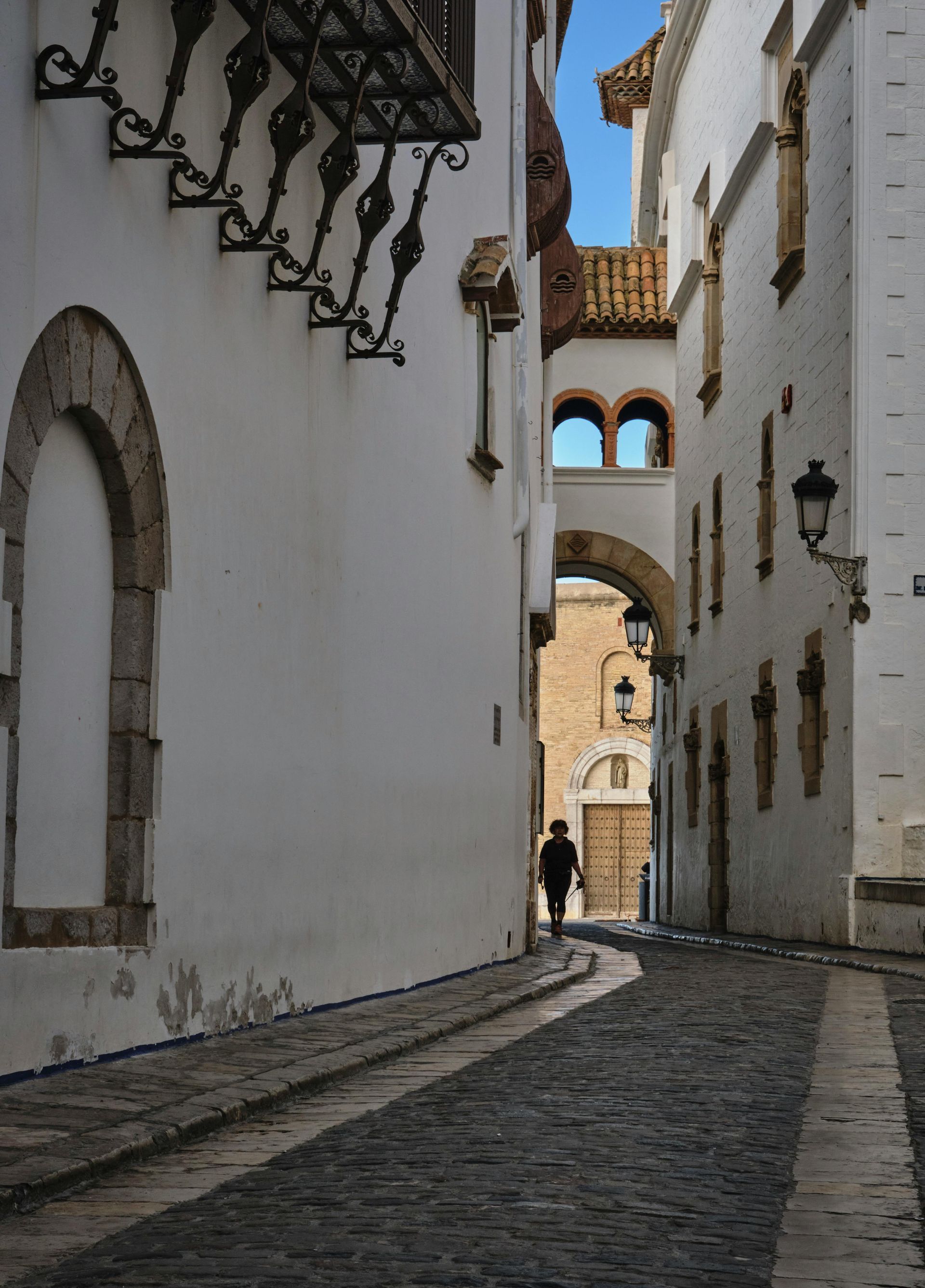 Narrow cobblestone street between white buildings, a person walks towards a distant archway.