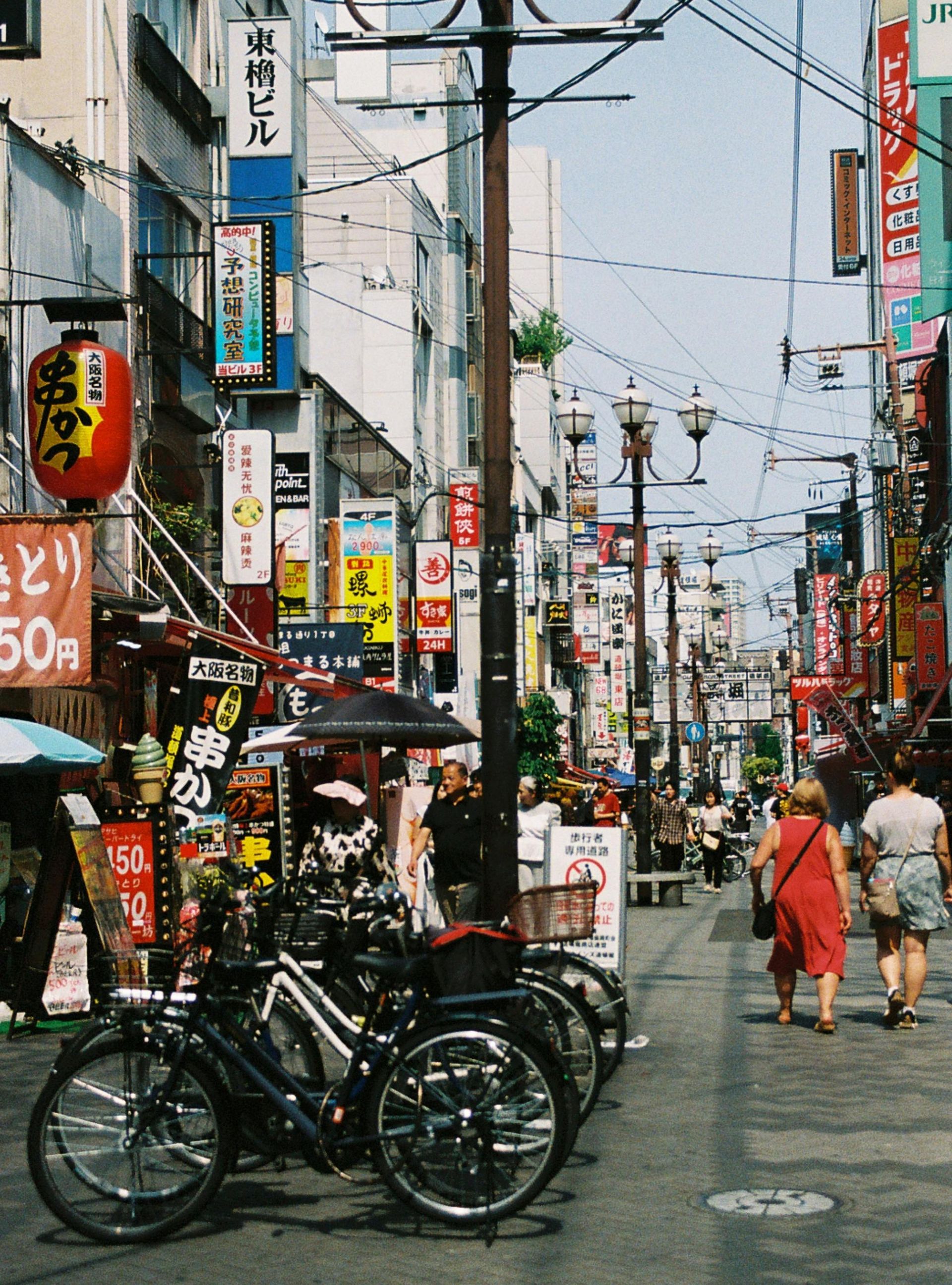 A sunny, narrow street in Japan filled with colorful storefront signs, a red lantern, and bicycles parked in the foreground.
