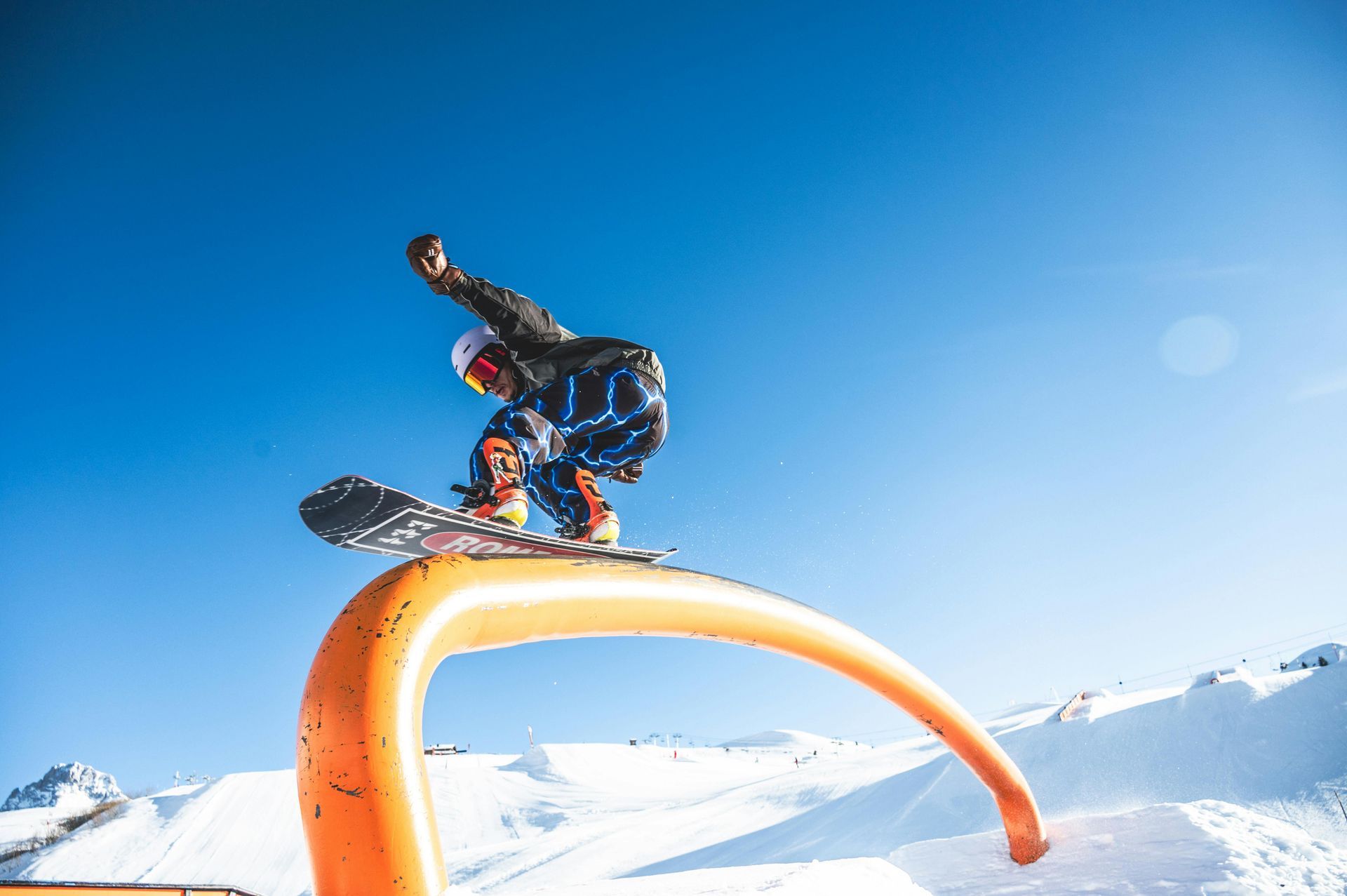 Snowboarder in mid-air performing a trick on an orange inflatable arch; blue sky and snowy mountains.