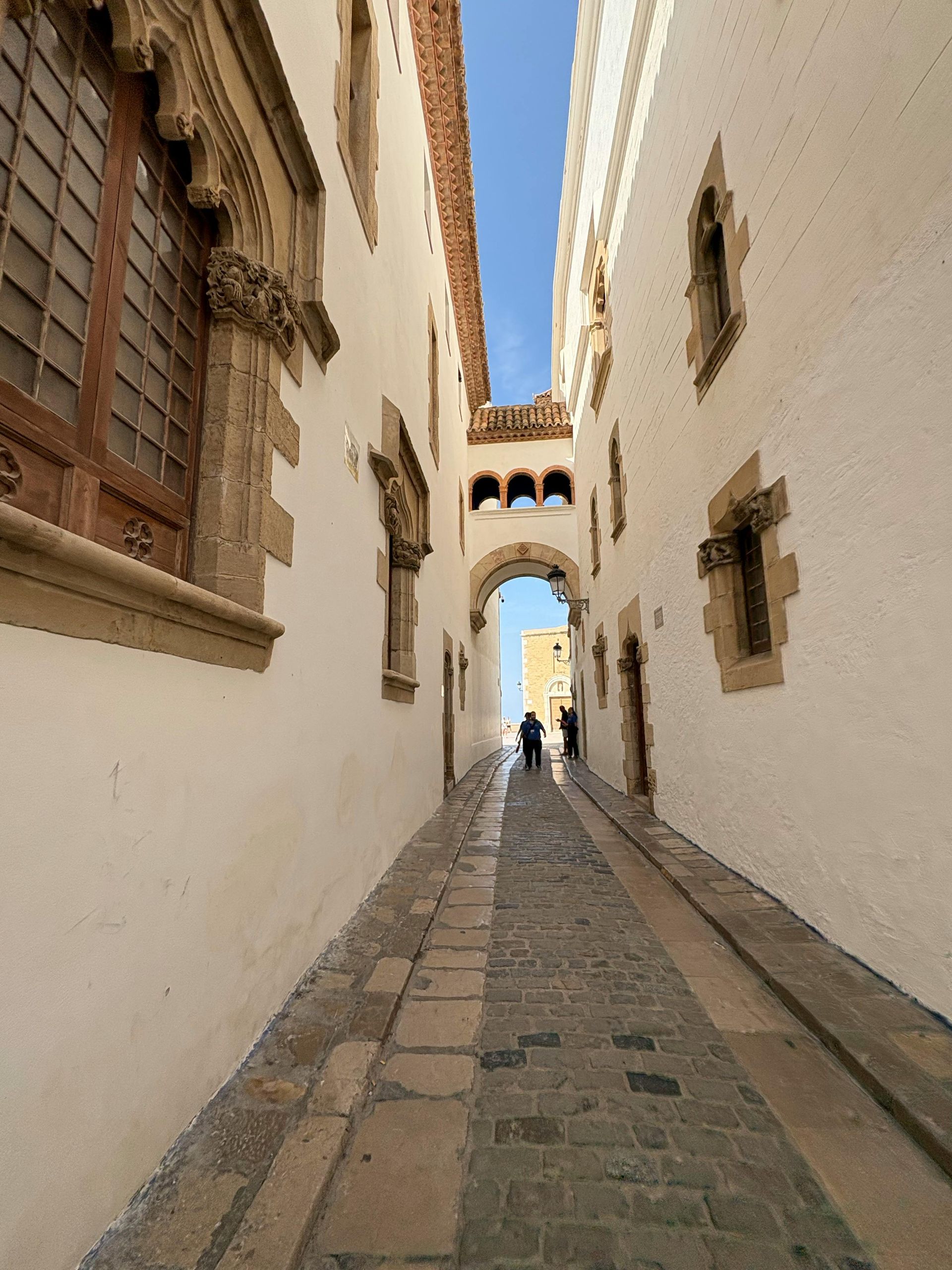 Narrow stone alleyway in a Spanish town, white buildings with arched openings, two figures walking towards the light. Sitges