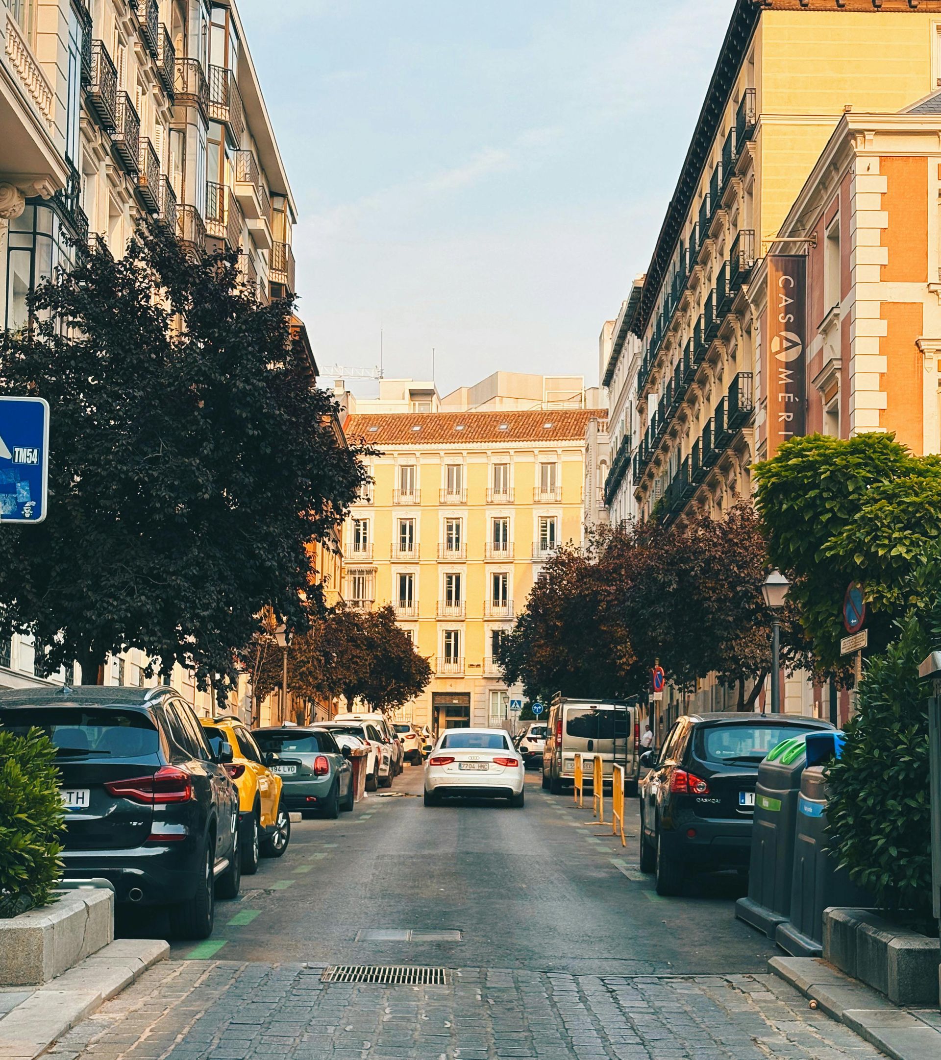 Street scene in Madrid with parked cars, buildings, trees, and a car driving towards the viewer. Chueca – Madrid