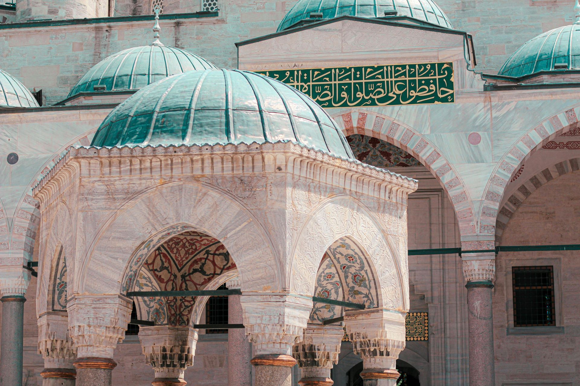 Ornate marble structure with a turquoise dome, part of a mosque in a sunlit setting.