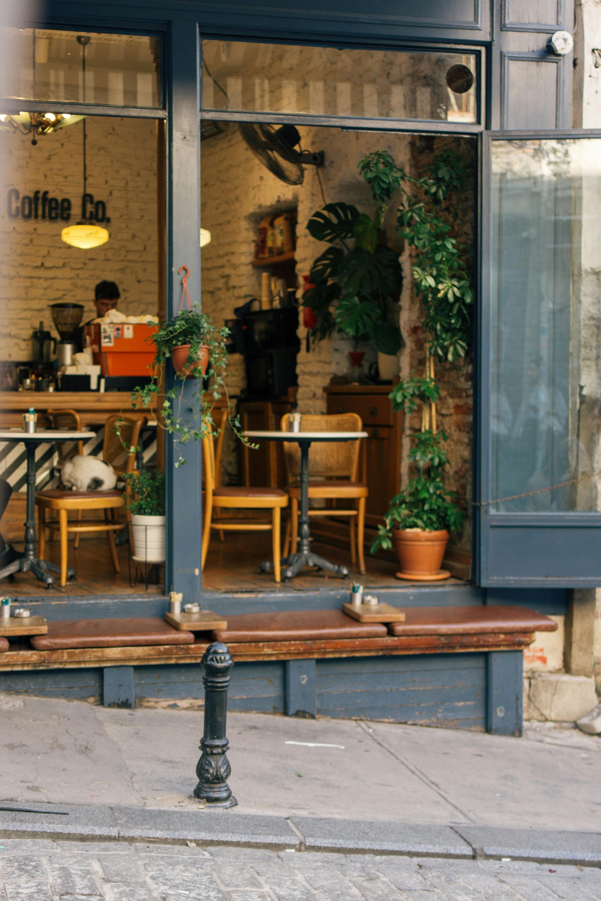 Small café storefront with wooden tables, potted plants, and a bench on the sidewalk