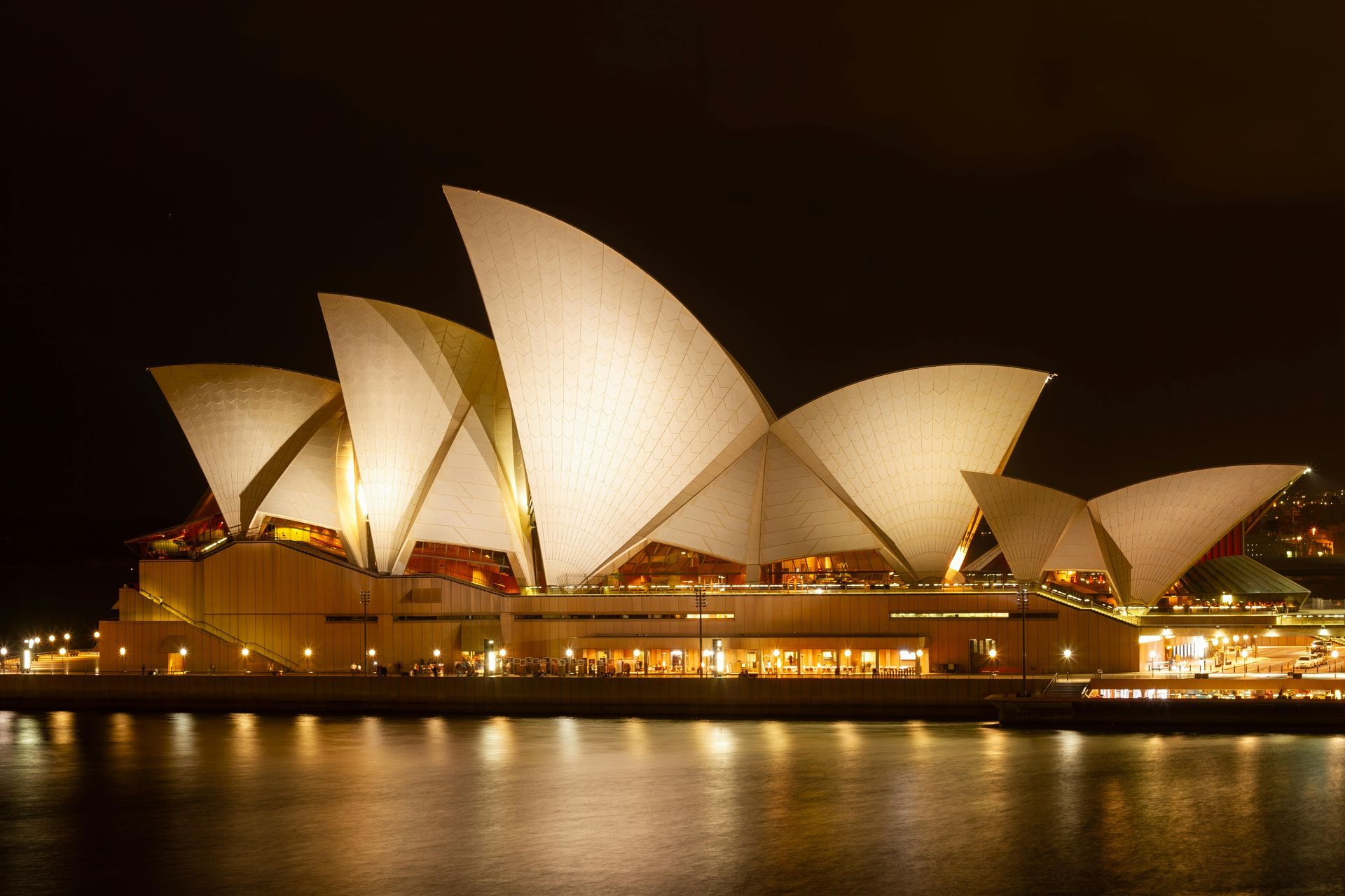 The Sydney Opera House illuminated at night, viewed from the harbor with its reflection on the water.