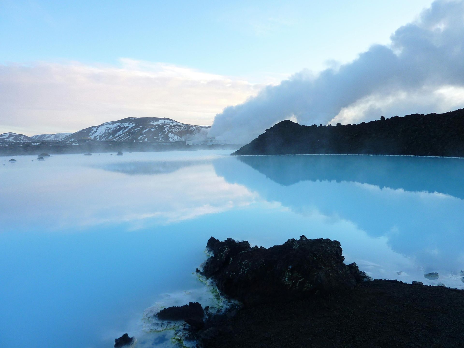 Blue geothermal lagoon with steam rising, dark rocks in foreground, snow-capped hills under a cloudy sky.