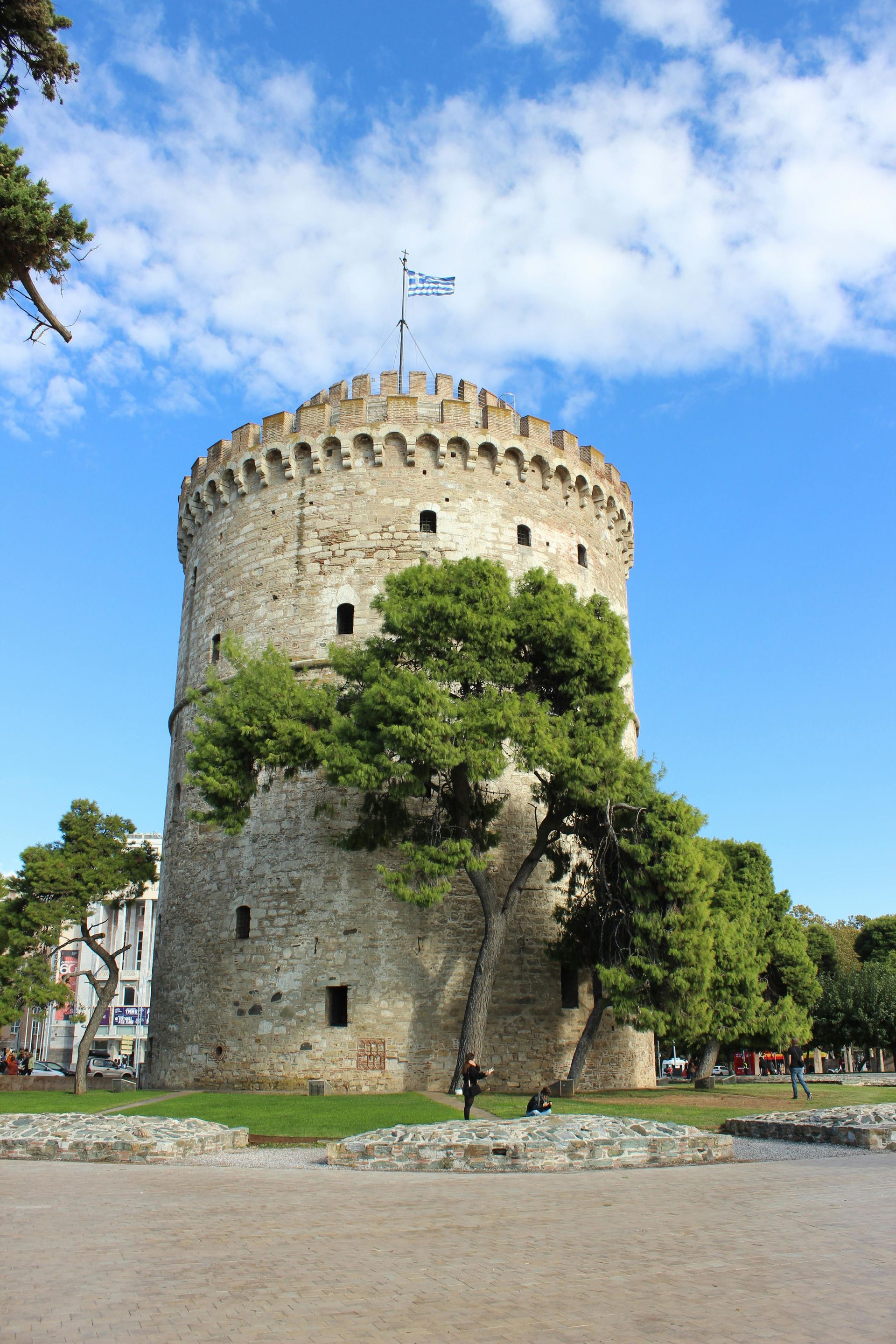 The White Tower of Thessaloniki, an ancient stone fortress, stands under a blue sky with trees in the foreground.