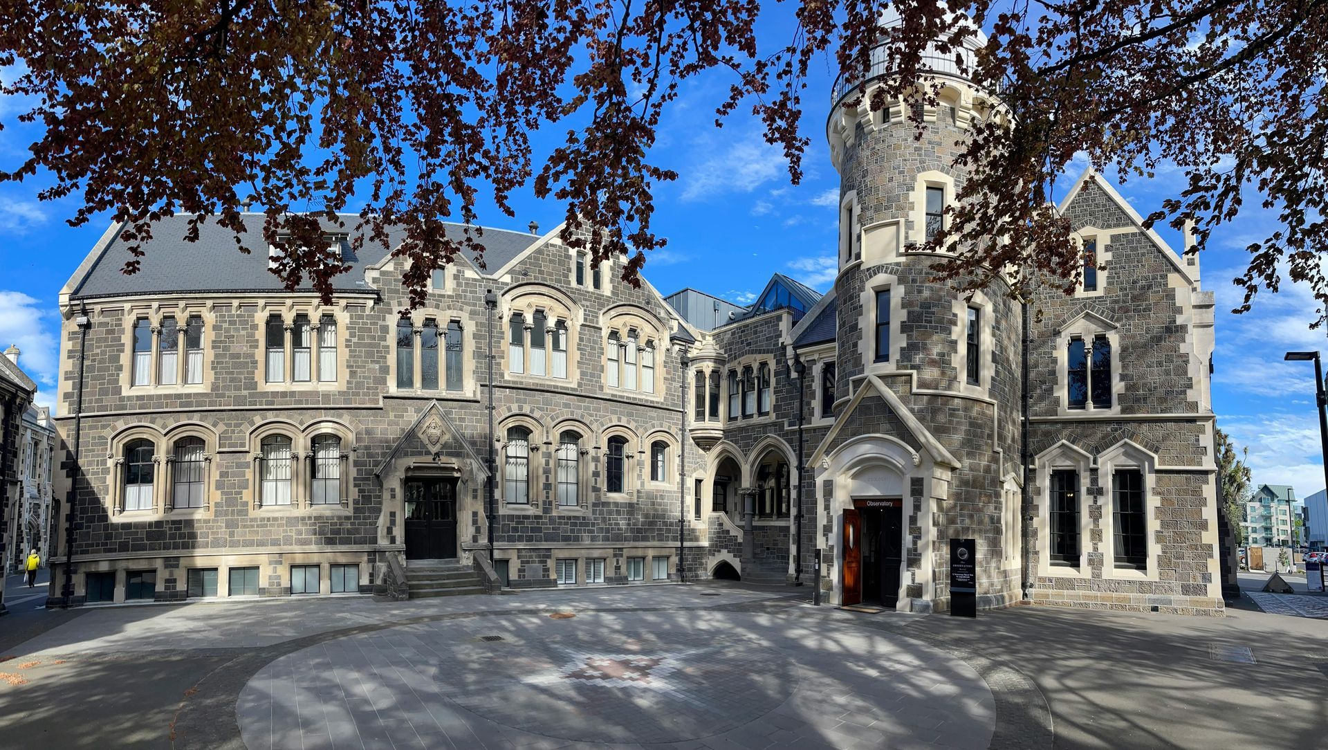 A historic stone building with a prominent turret, viewed across a paved courtyard under a bright blue sky.