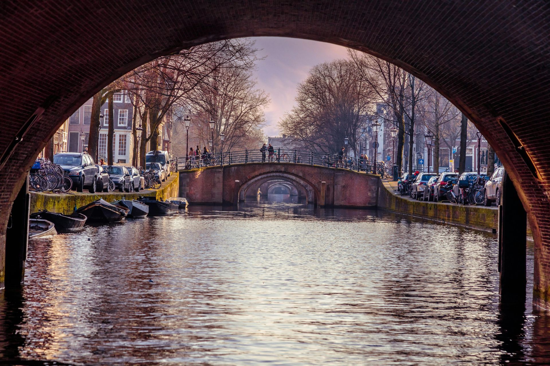 A canal in Amsterdam framed by a brick archway, showing bridges, parked cars, and bare trees under soft lighting.