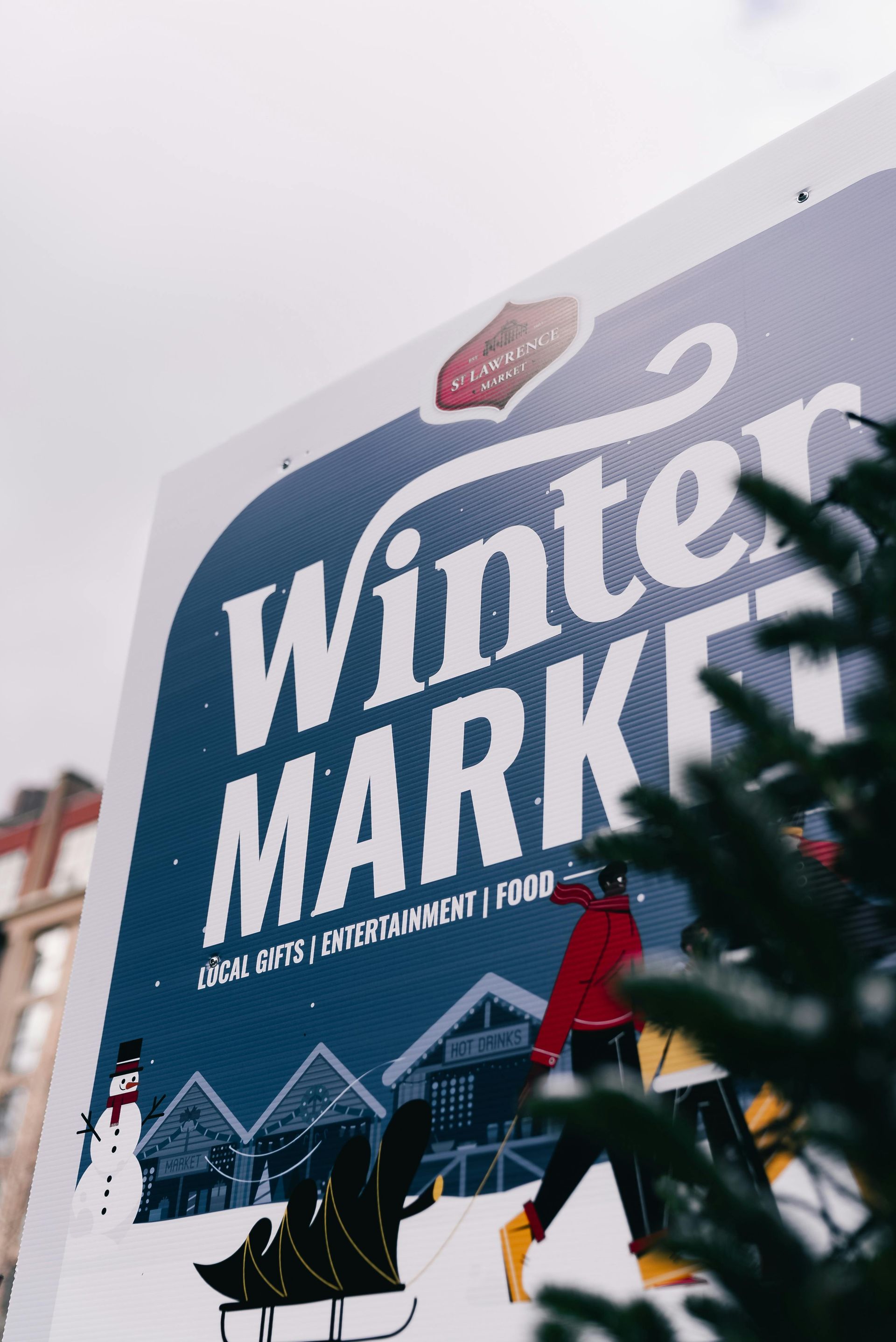 Winter Market sign with cartoon figures, snow, and a Christmas tree in the foreground.