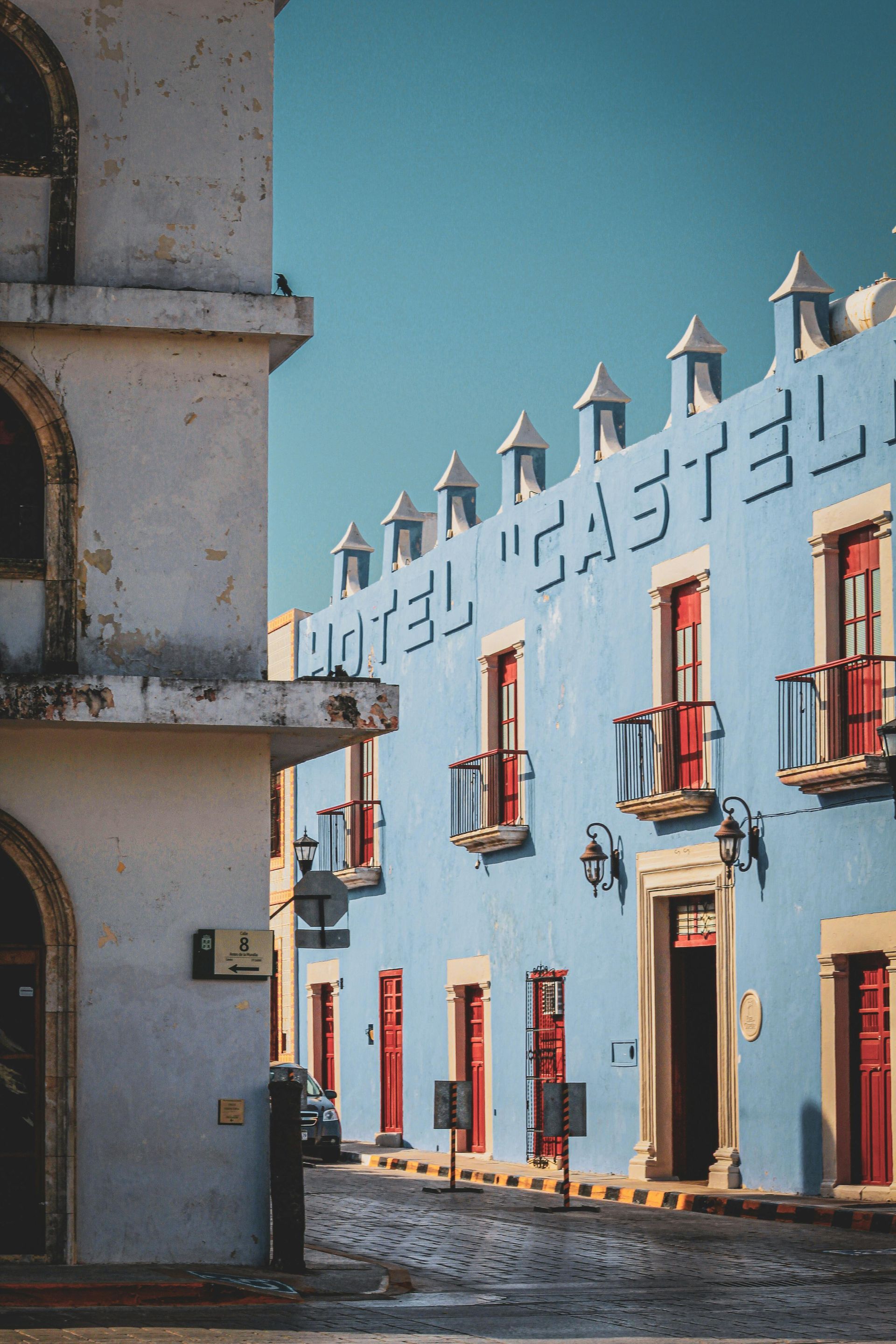 Buildings on a cobblestone street. A blue building has red doors and windows. A white building is to the left. Sunny day.