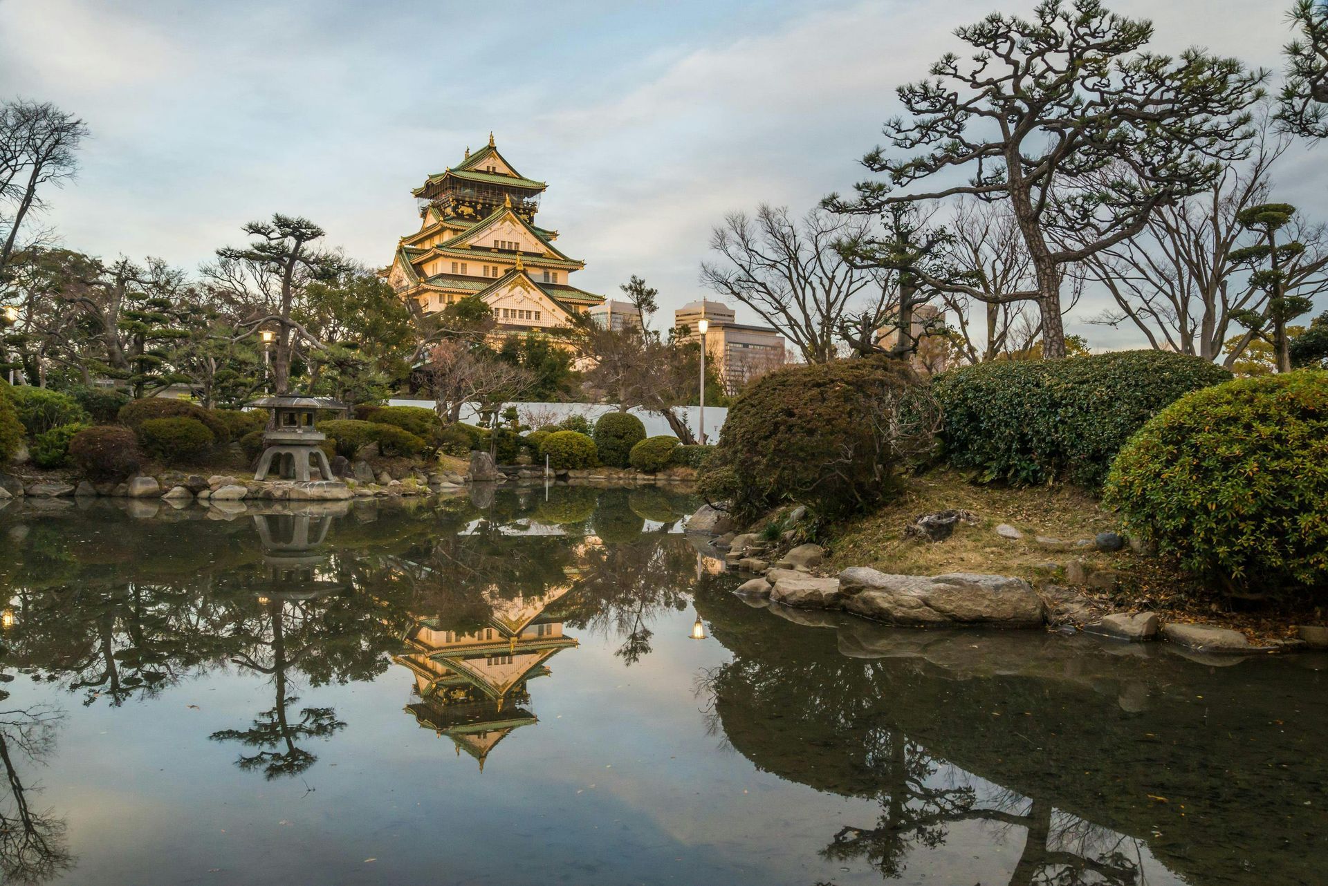 Japanese castle reflected in a still pond, framed by trees and a cloudy sky.