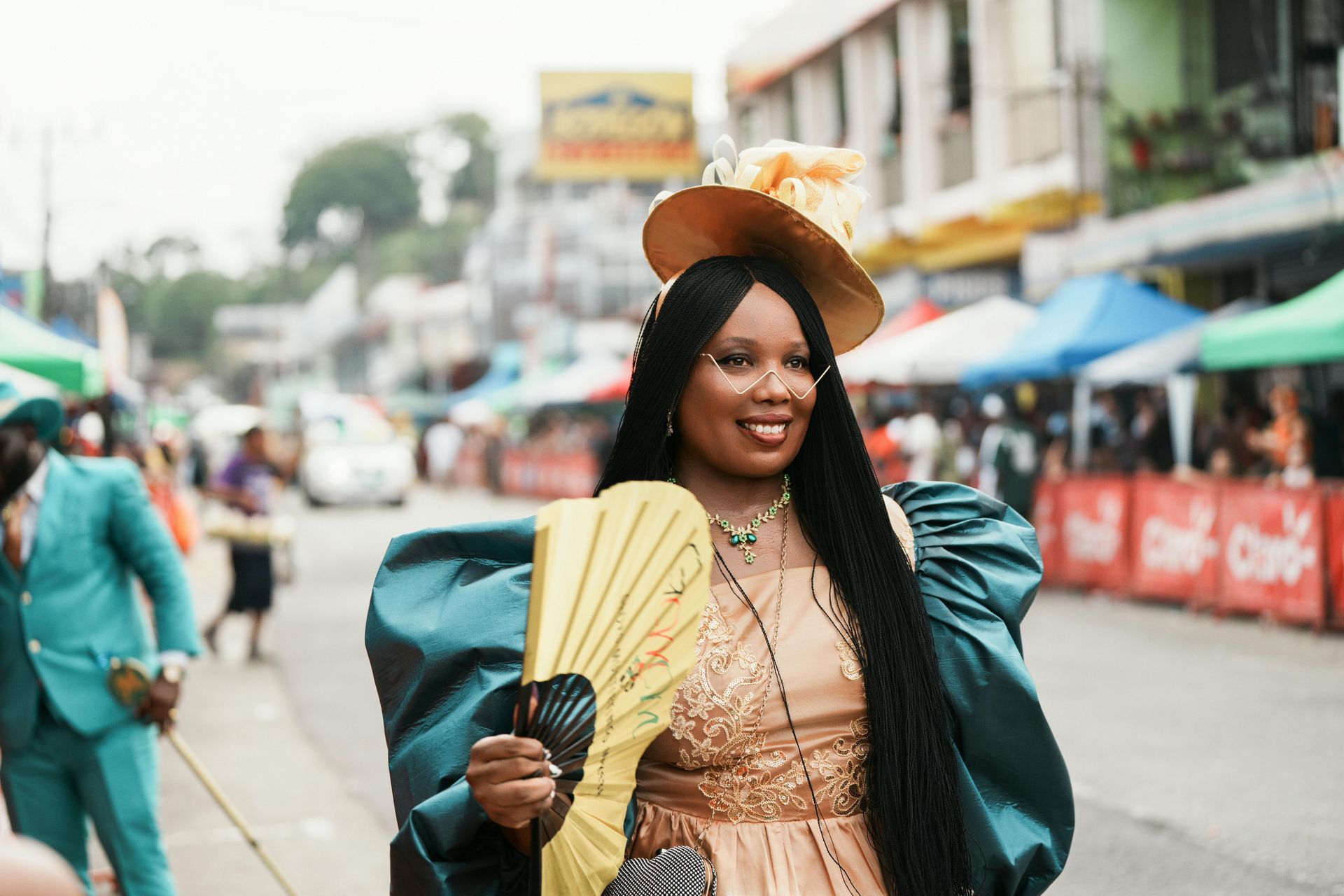 Woman in ornate costume, holding a fan, smiling. Street festival with colorful stalls and people in the background.