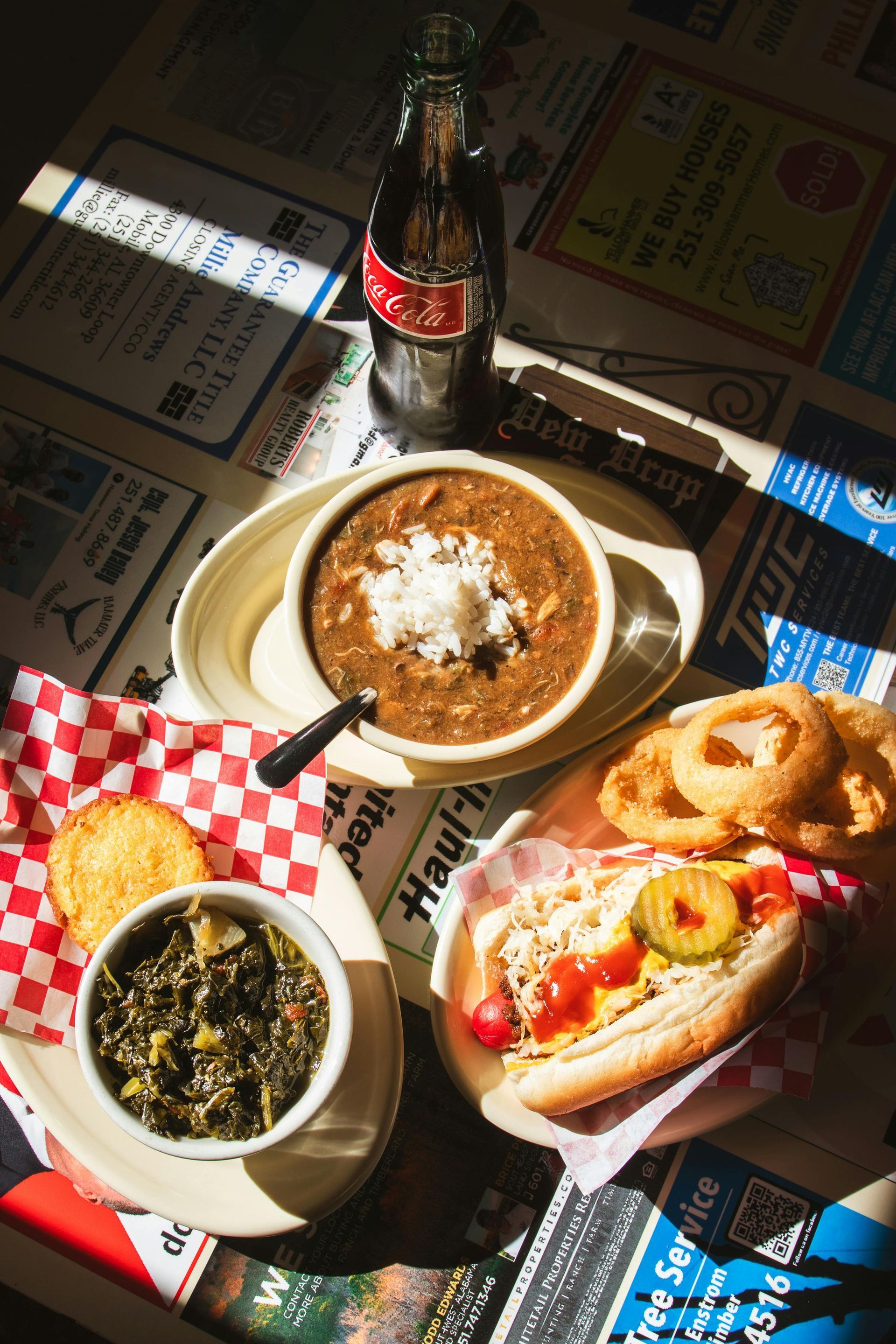 A top-down view of a diner table with chili, a hot dog, onion rings, a side of greens, and a glass bottle of Coca-Cola.