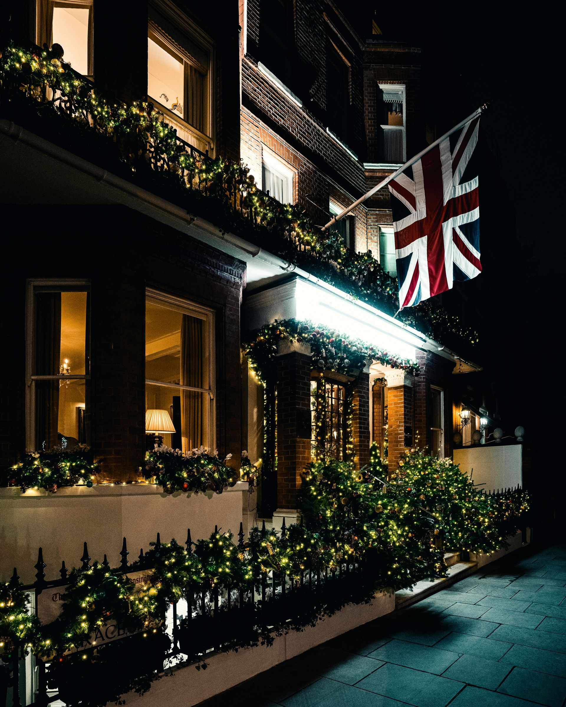 A brick building with a Union Jack flag at night, illuminated by lights and greenery.