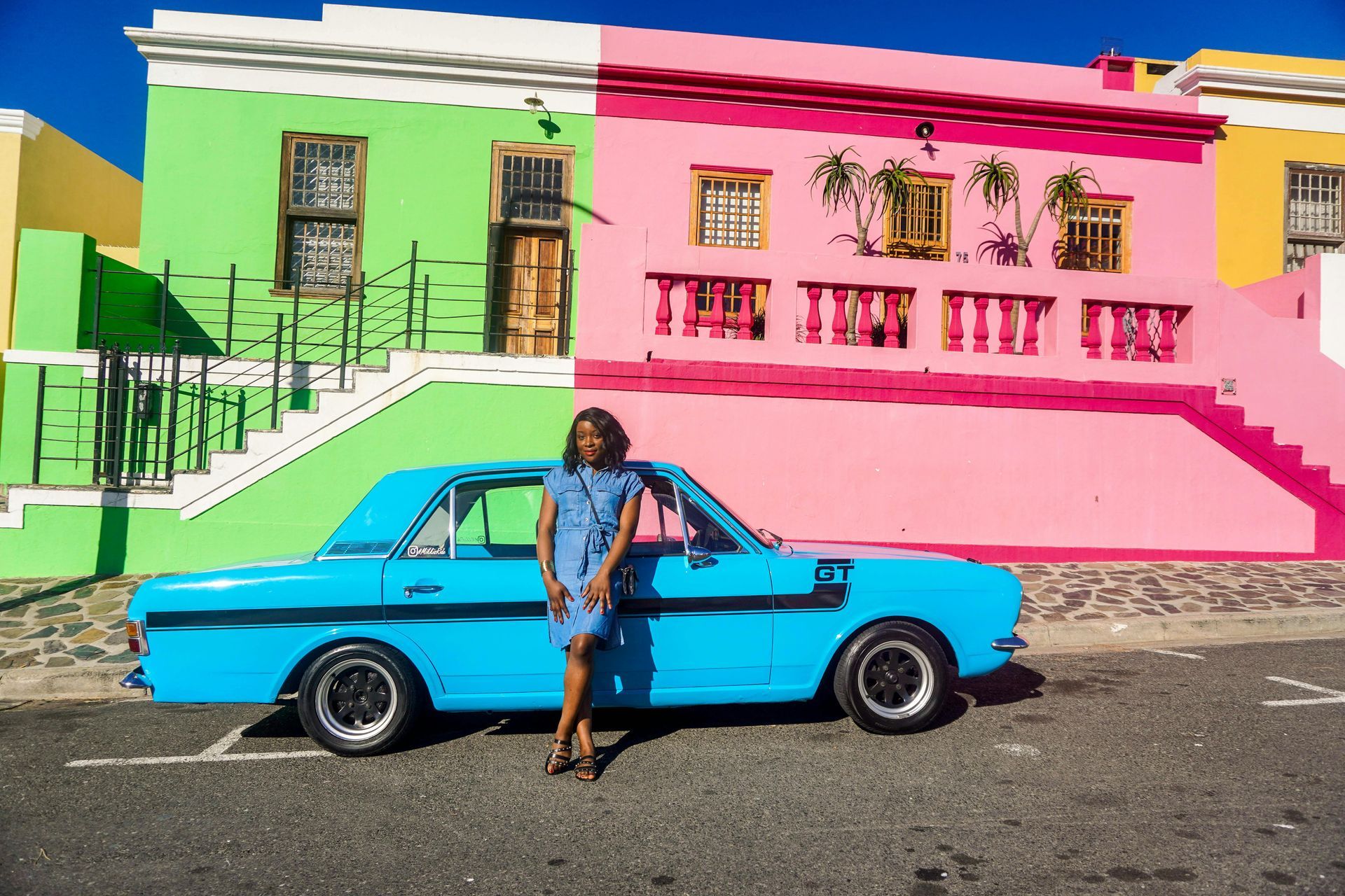 Woman leaning on a turquoise car in front of a brightly painted pink, green, and yellow building.