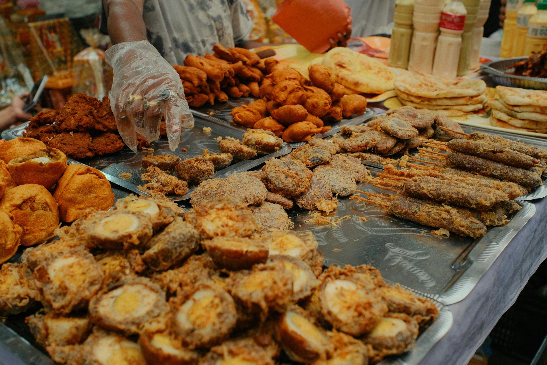 A person wearing a plastic glove selects fried foods, including breaded eggs and pastries, from trays at a market stand.