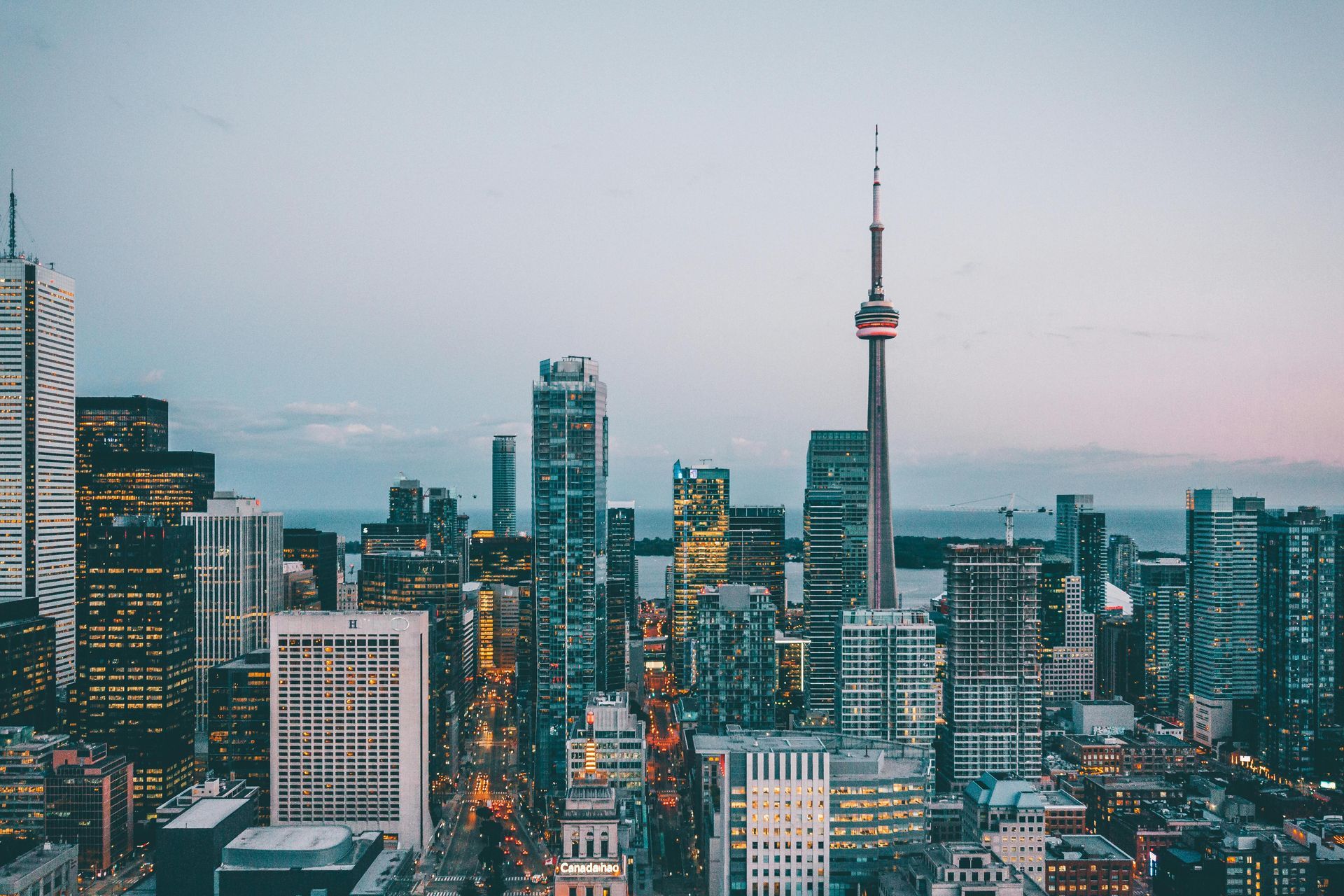 Toronto skyline at dusk, with the CN Tower prominent. Buildings lit up, a body of water in the background.