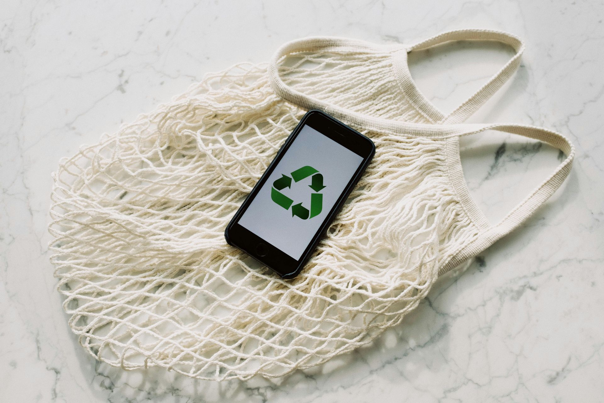 A smartphone with a green recycling symbol on its screen rests inside a white, woven mesh shopping bag on a marble surface.