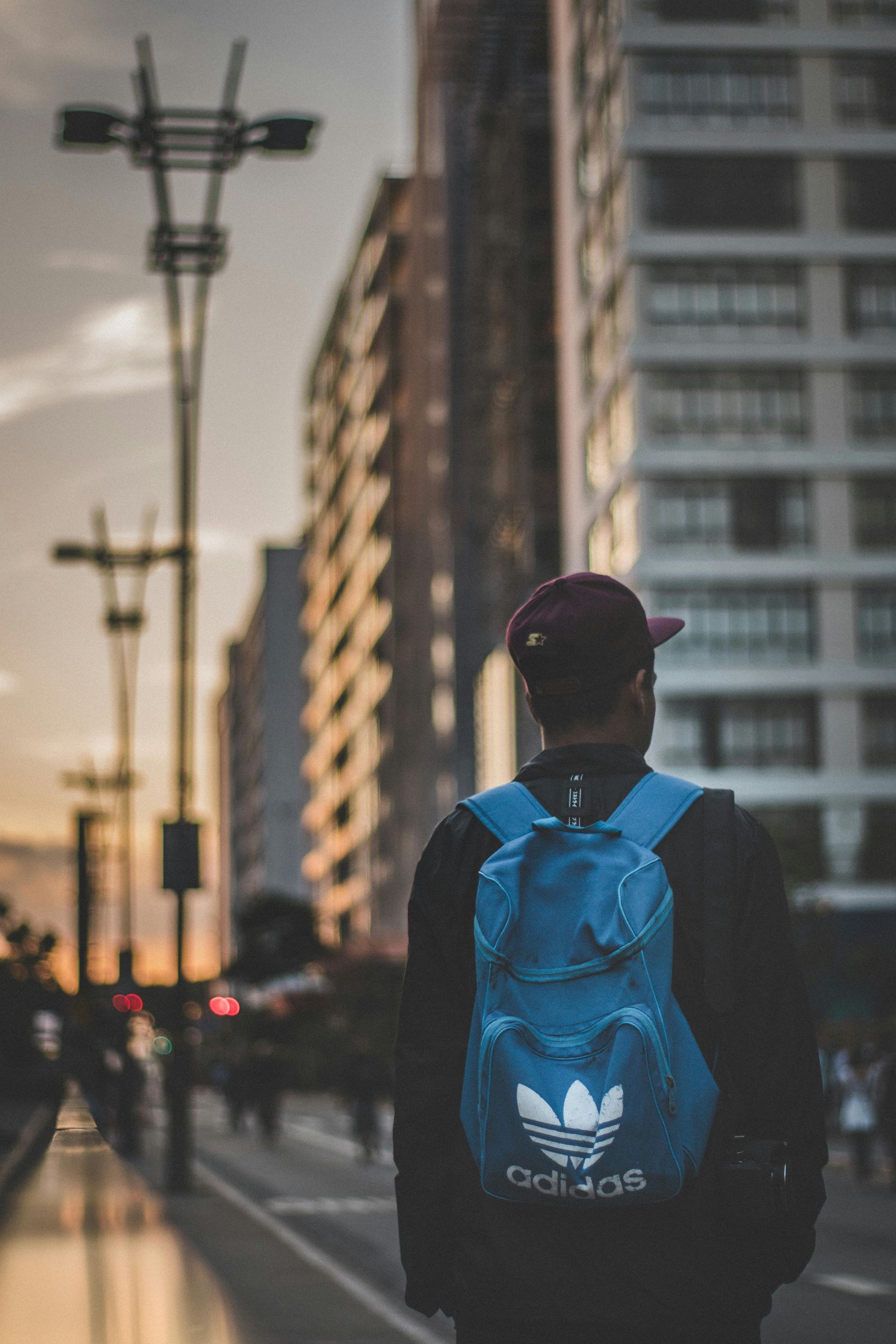 Person with blue backpack and Adidas logo walking in a city, buildings in background, dusk.
