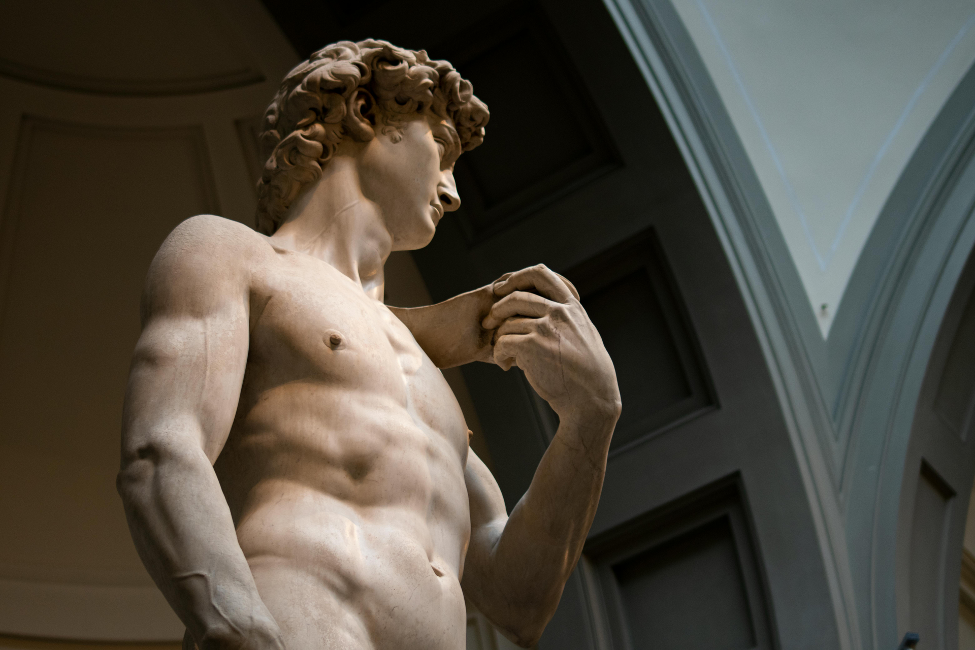 Michelangelo's statue of David, a muscular nude male figure, looking to the side, set in a gallery.