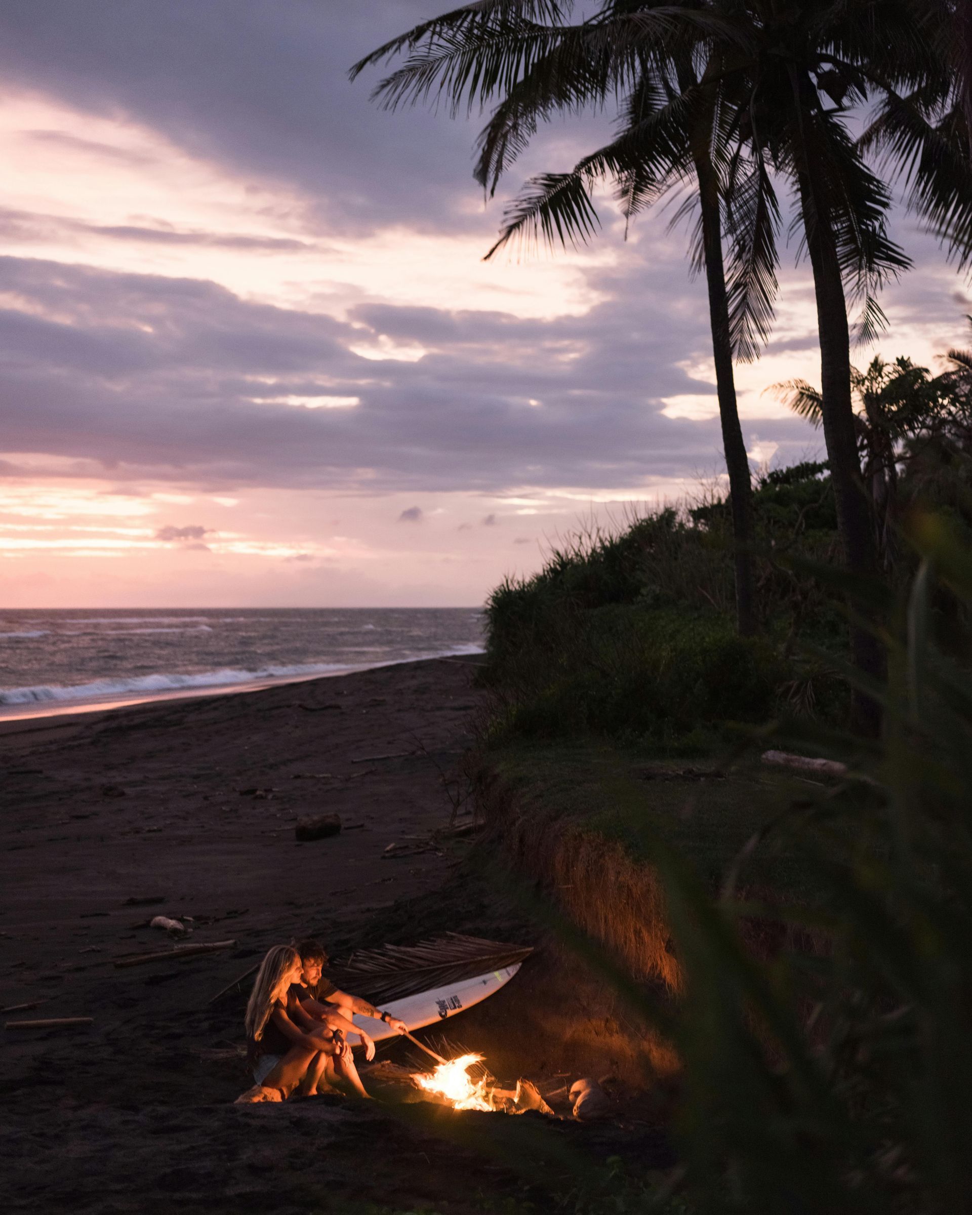 Beach bonfire at dusk: waves, dark sand, palm trees, and a surfboard by the fire under a colorful, cloudy sky. Fire Island, New York,