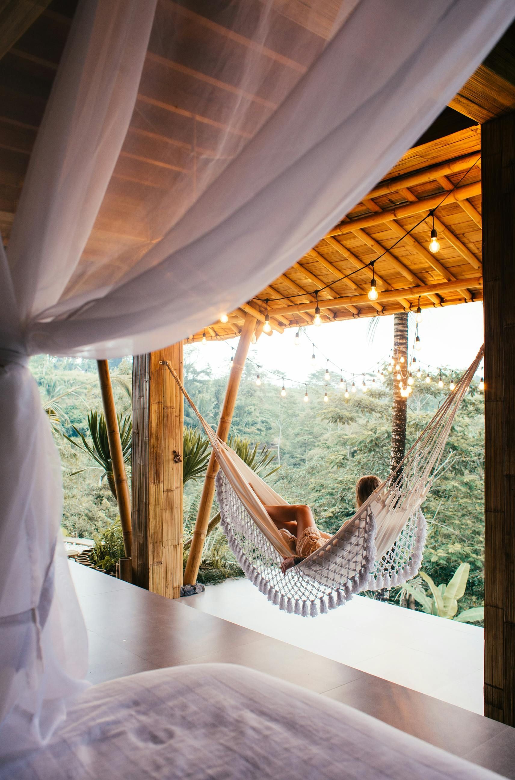 A person relaxes in a white crocheted hammock overlooking a lush, forested valley from a thatched-roof balcony.