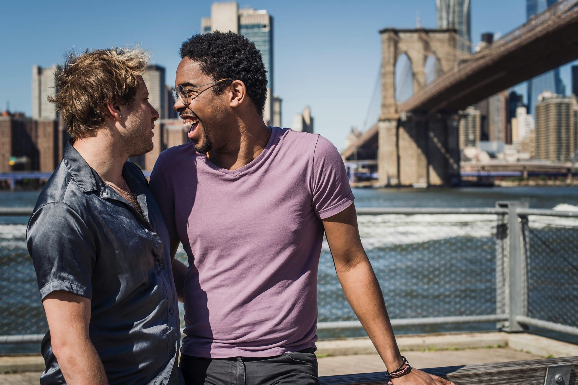 Two men, smiling, by water, Brooklyn Bridge in background.