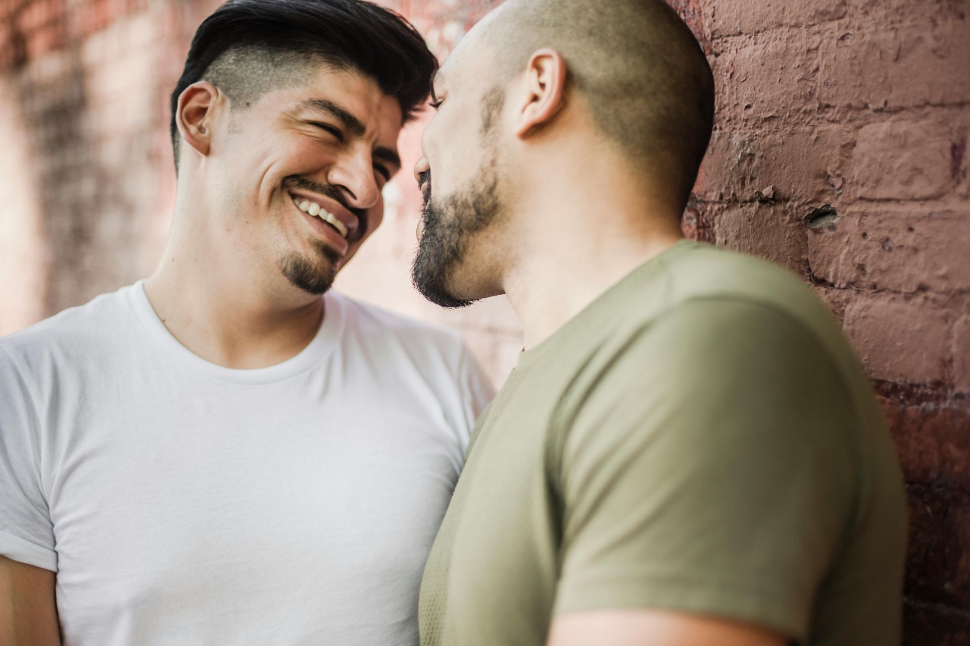 Two men smiling at each other, leaning against a brick wall. One wears white, the other olive.
