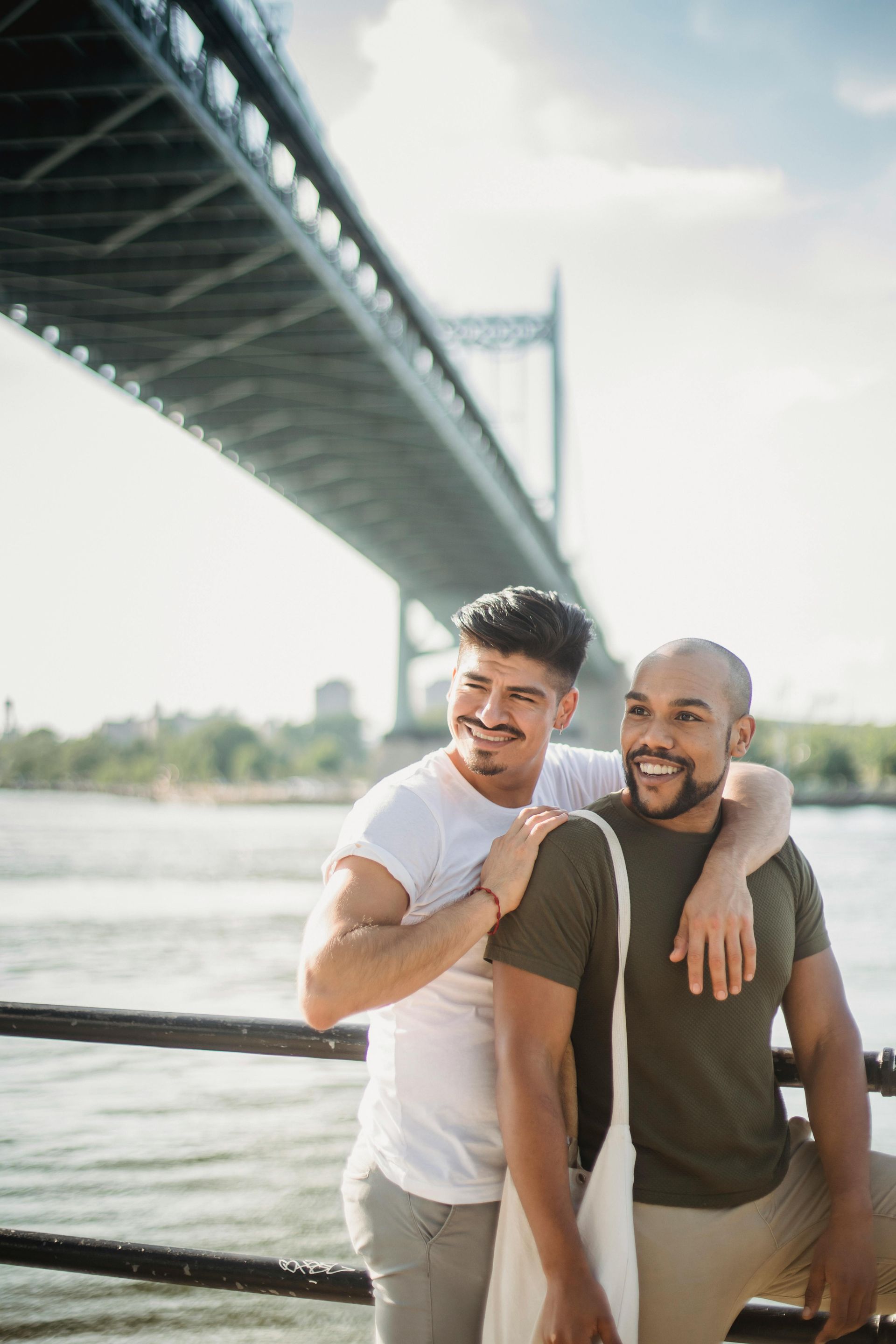 Two men smiling by a riverside railing under a large bridge