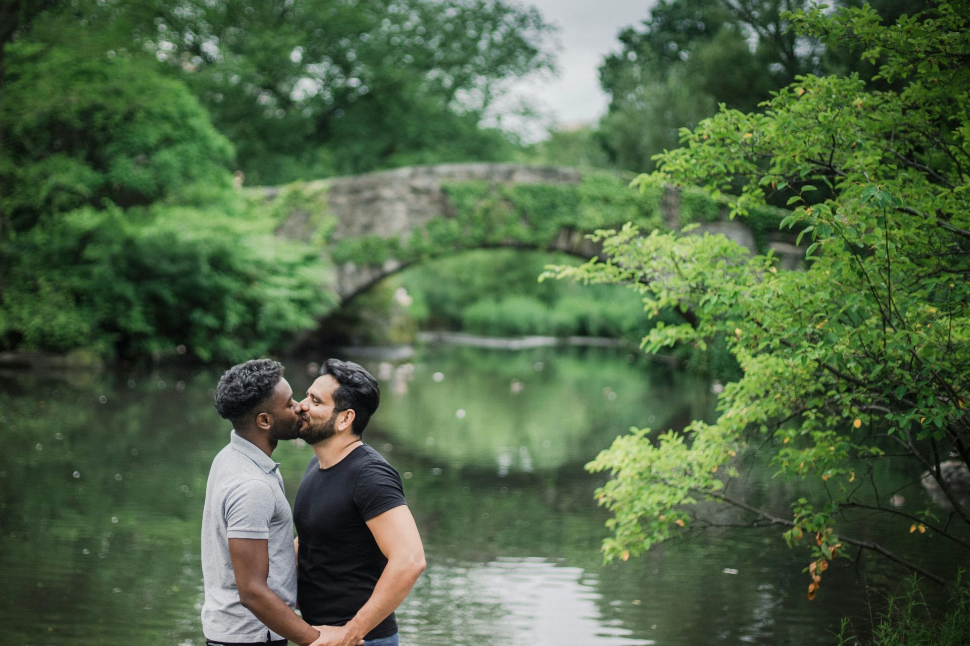 Two men kissing by a river with a stone bridge in the background, surrounded by lush greenery.