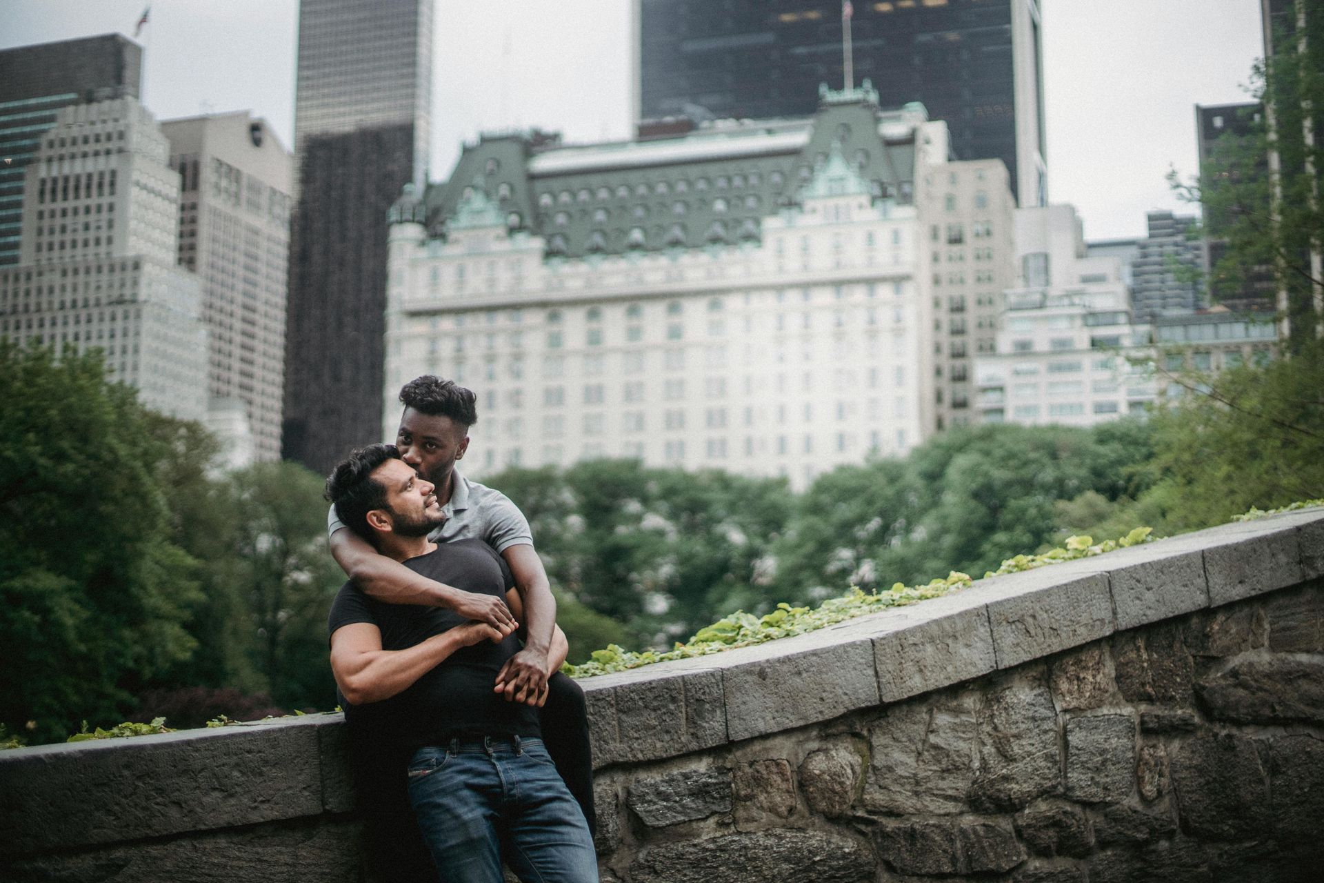 Two men embrace outdoors in New York City, against a stone wall with skyscrapers in background.