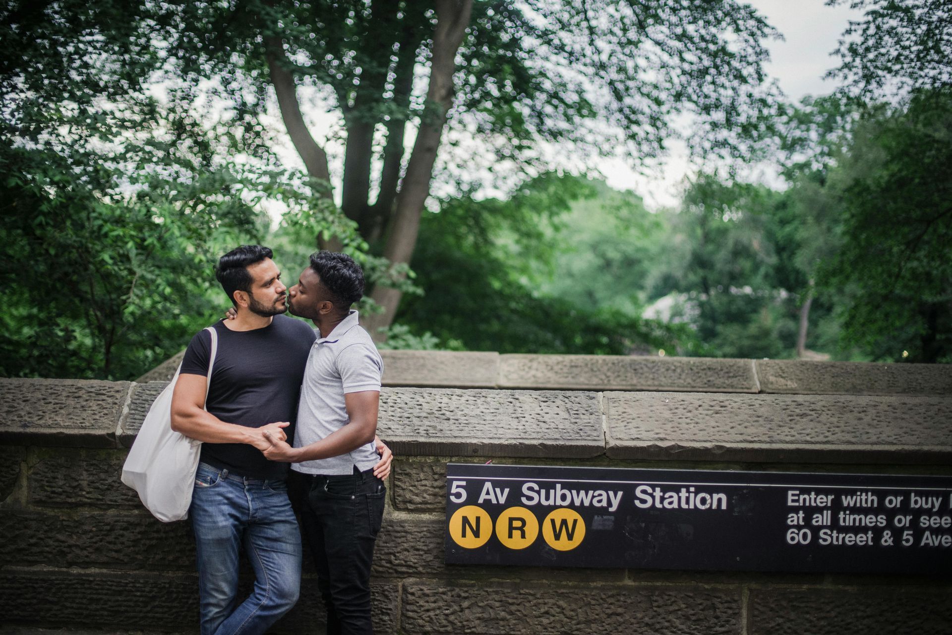 Two men embrace at a subway station. One is kissing the other's cheek; a stone wall and trees are behind them.