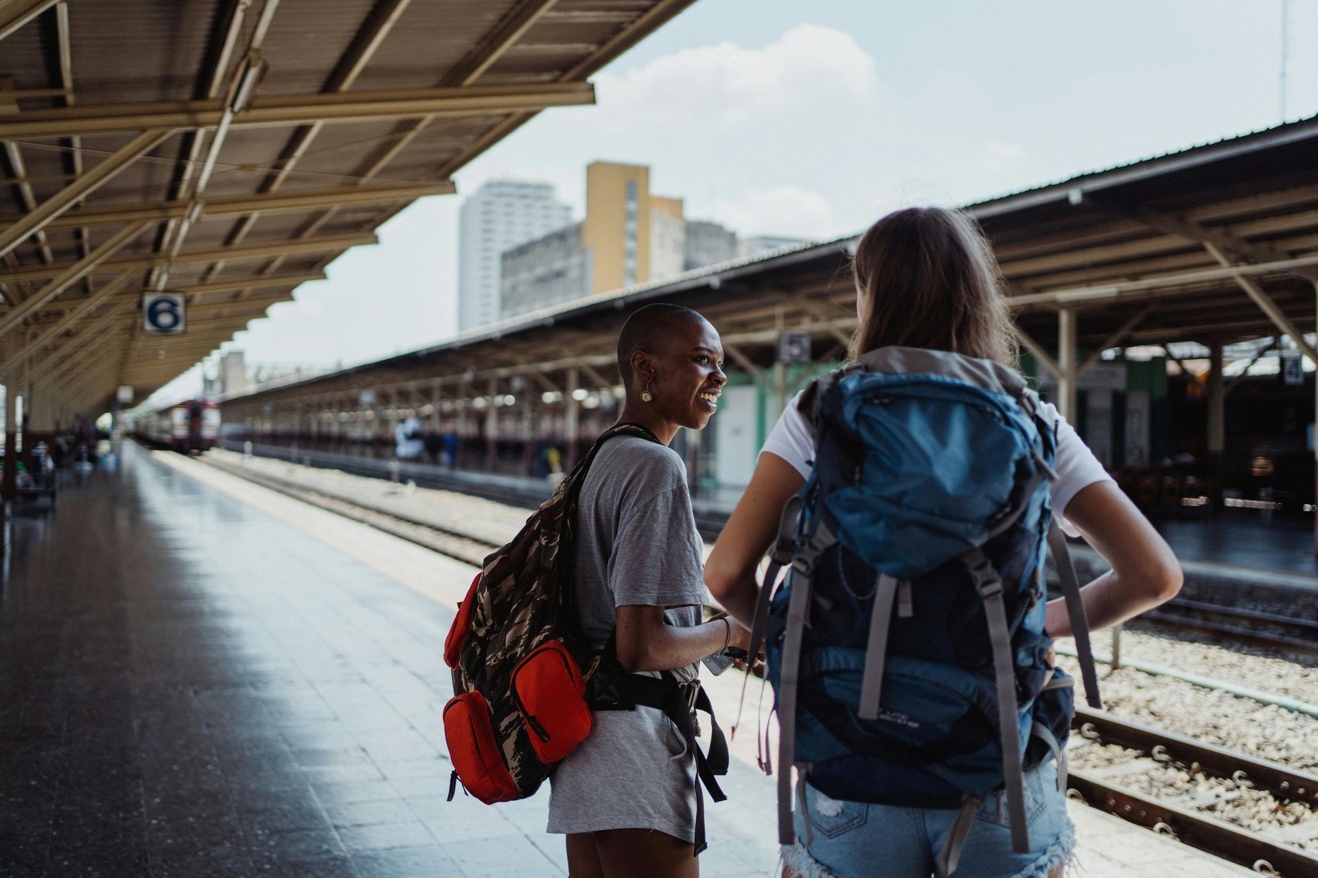 Two women with backpacks at a train station, smiling and chatting, tracks visible in background.