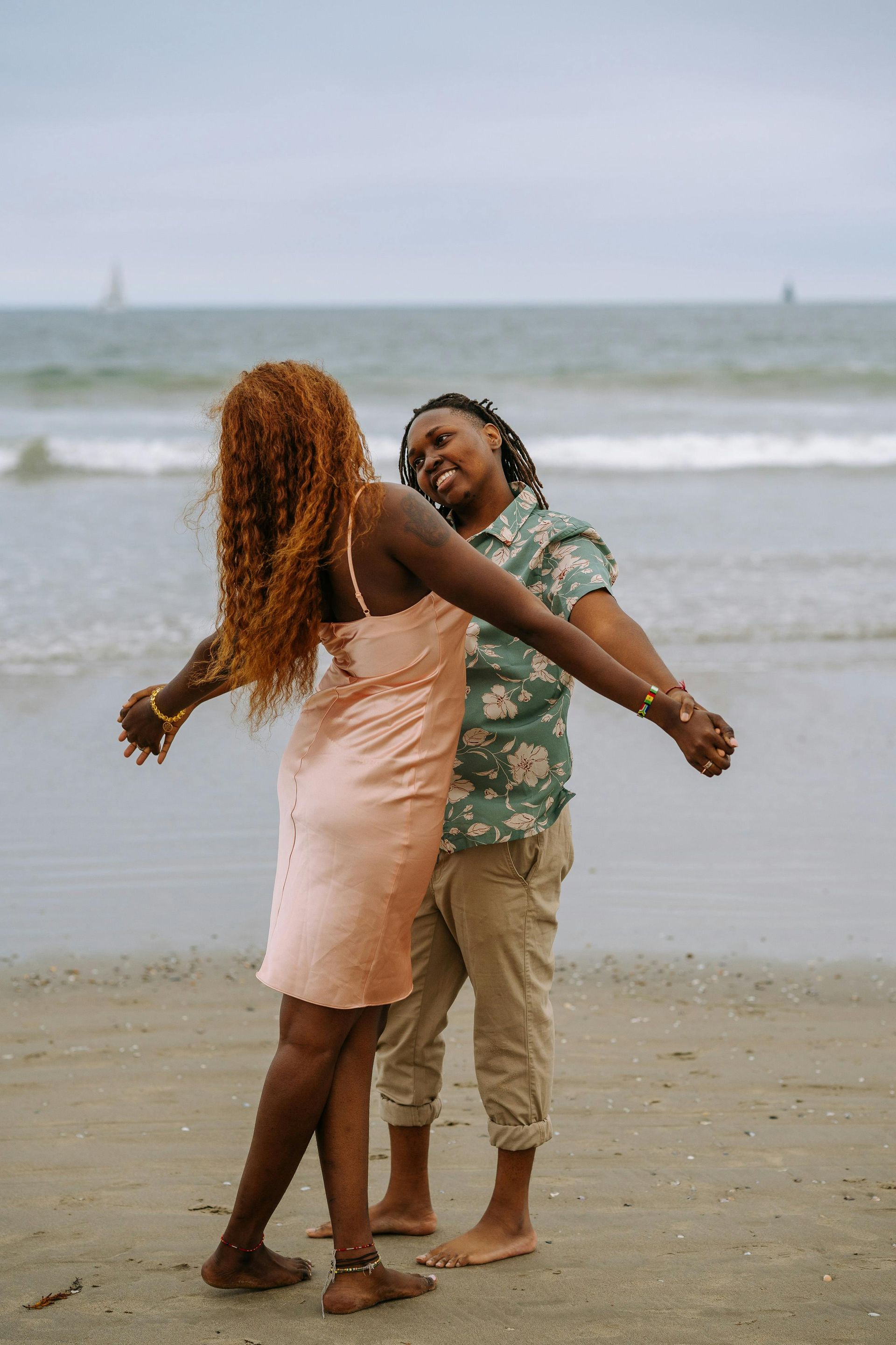 Two people embracing on a beach, smiling. One in a pink dress, other in a green shirt and khaki pants. Ocean in background. LESBIANS!!