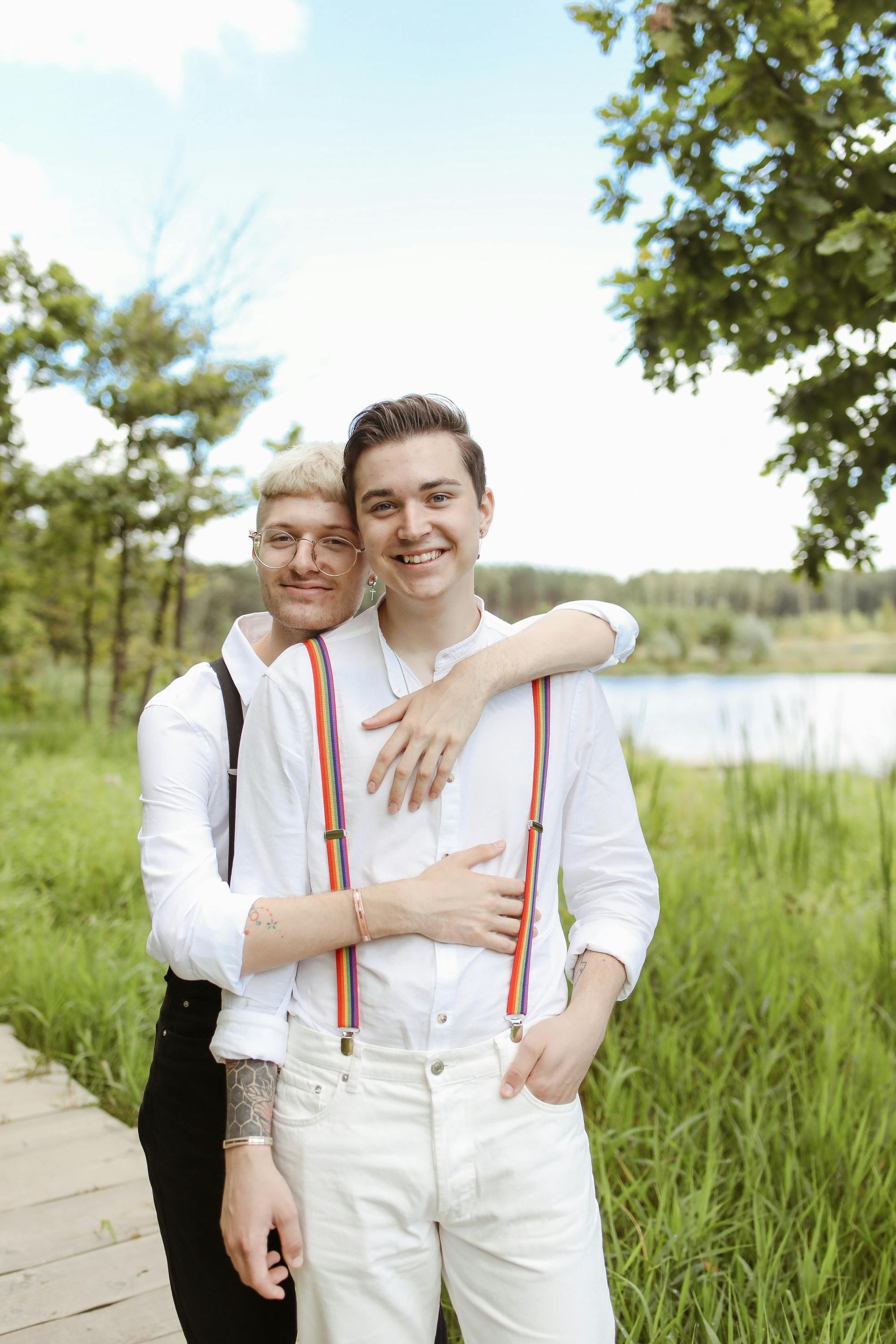 Two people in white shirts embrace on a boardwalk near a grassy field and a lake.