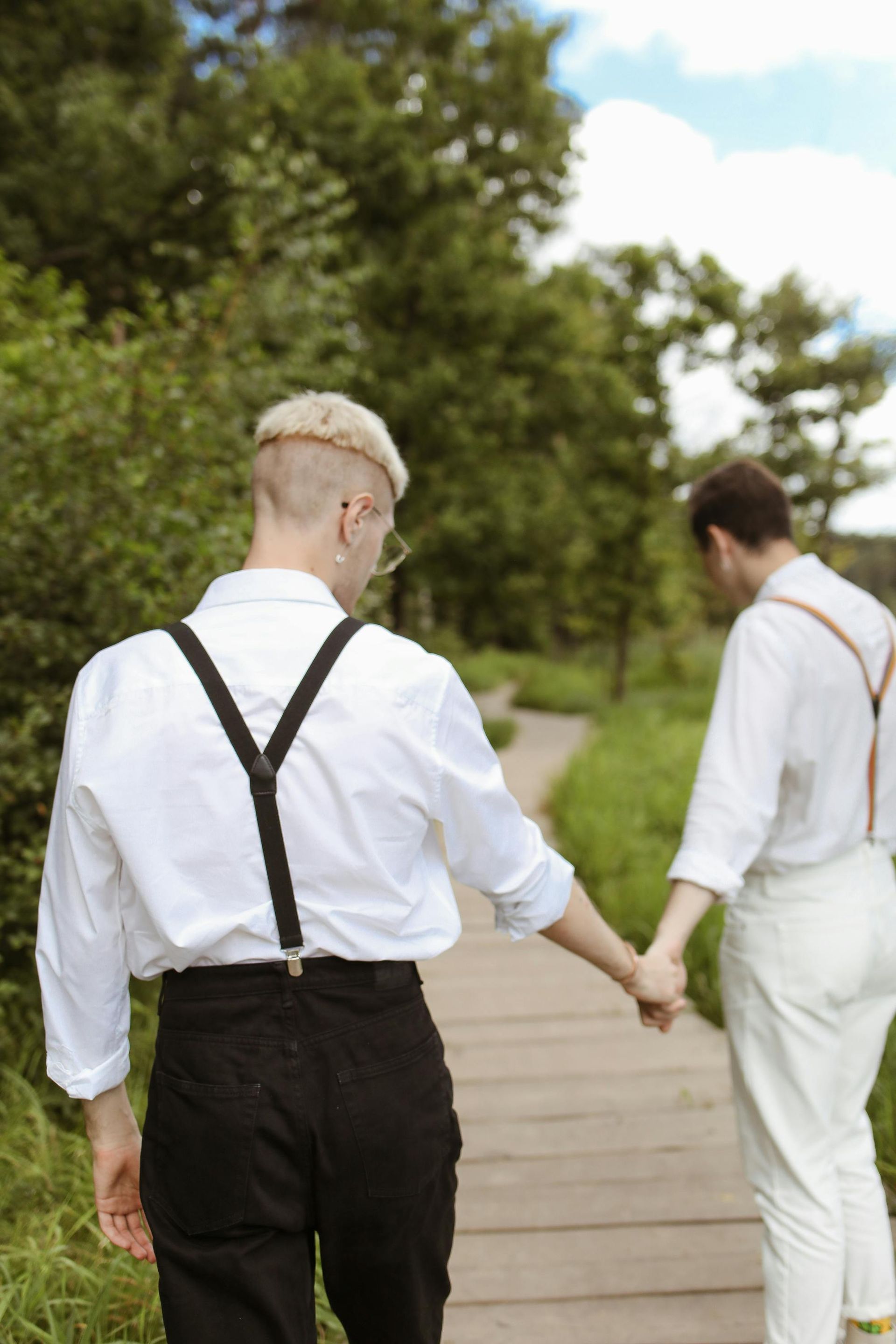 Two people hold hands and walk on a wooden path surrounded by greenery. One wears black pants, the other, white pants and suspenders.