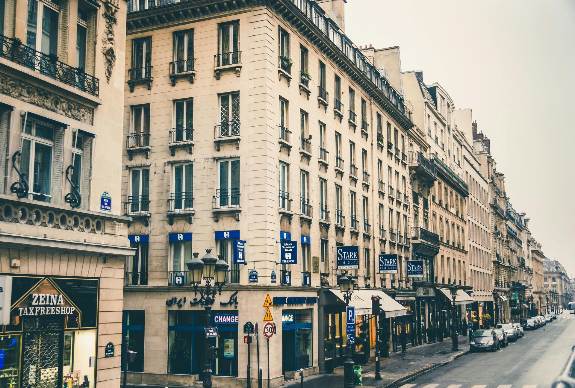 Buildings line a street in Paris, France; shops and cars are visible. Overcast sky.