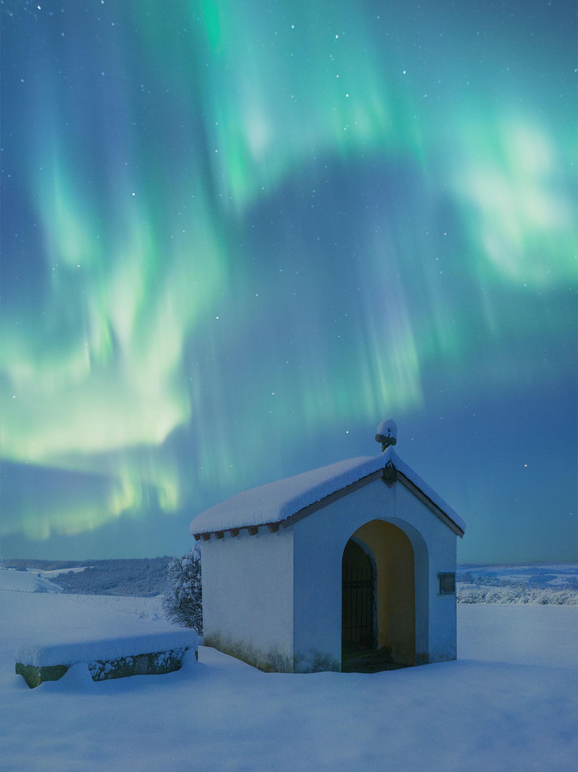 Chapel under green and blue aurora borealis in a snowy landscape.