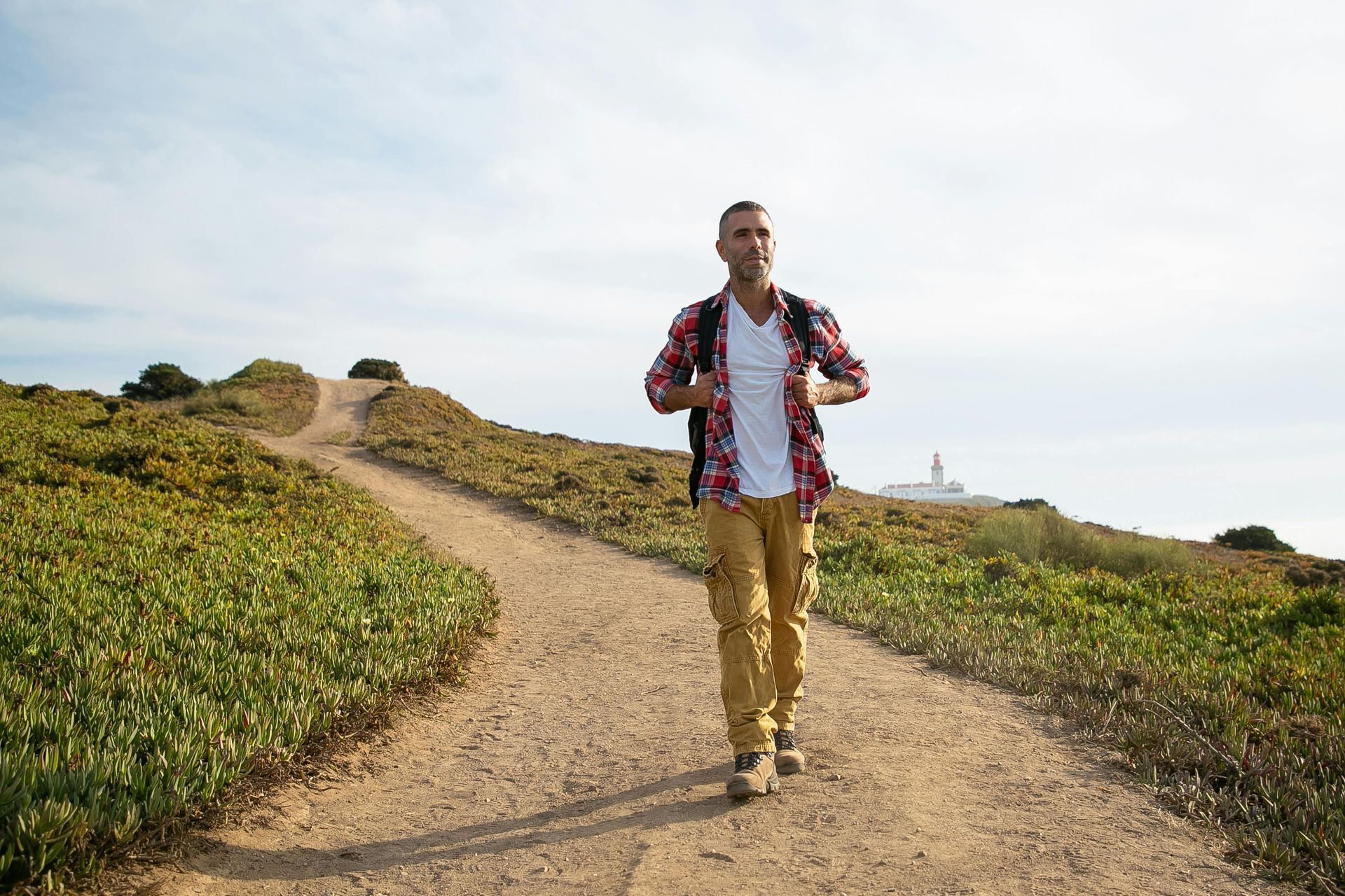 Man hiking on a dirt path wearing a backpack, plaid shirt, and khakis, near green plants under a blue sky.
