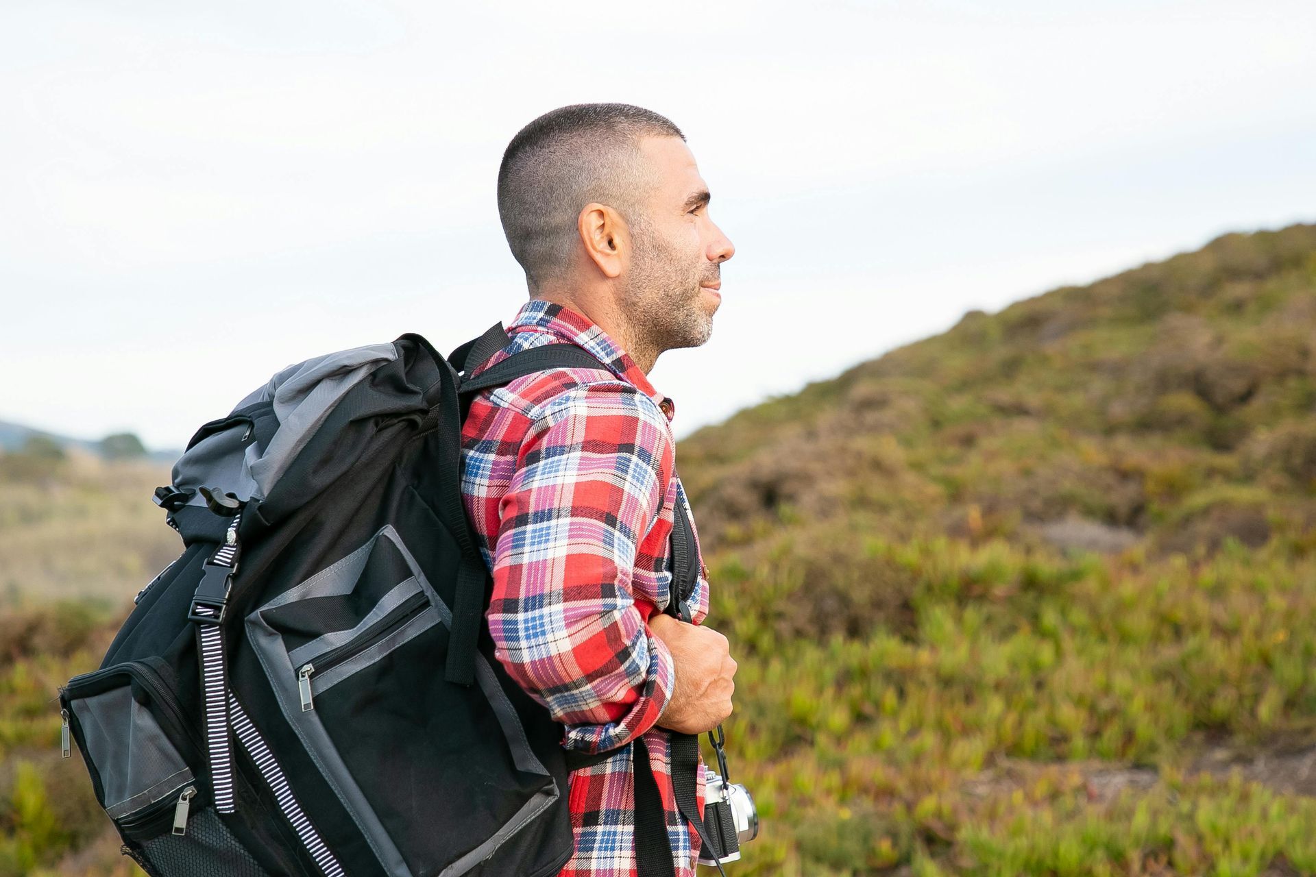 Hiker in a red plaid shirt with a large black backpack, standing outdoors on a grassy hillside.