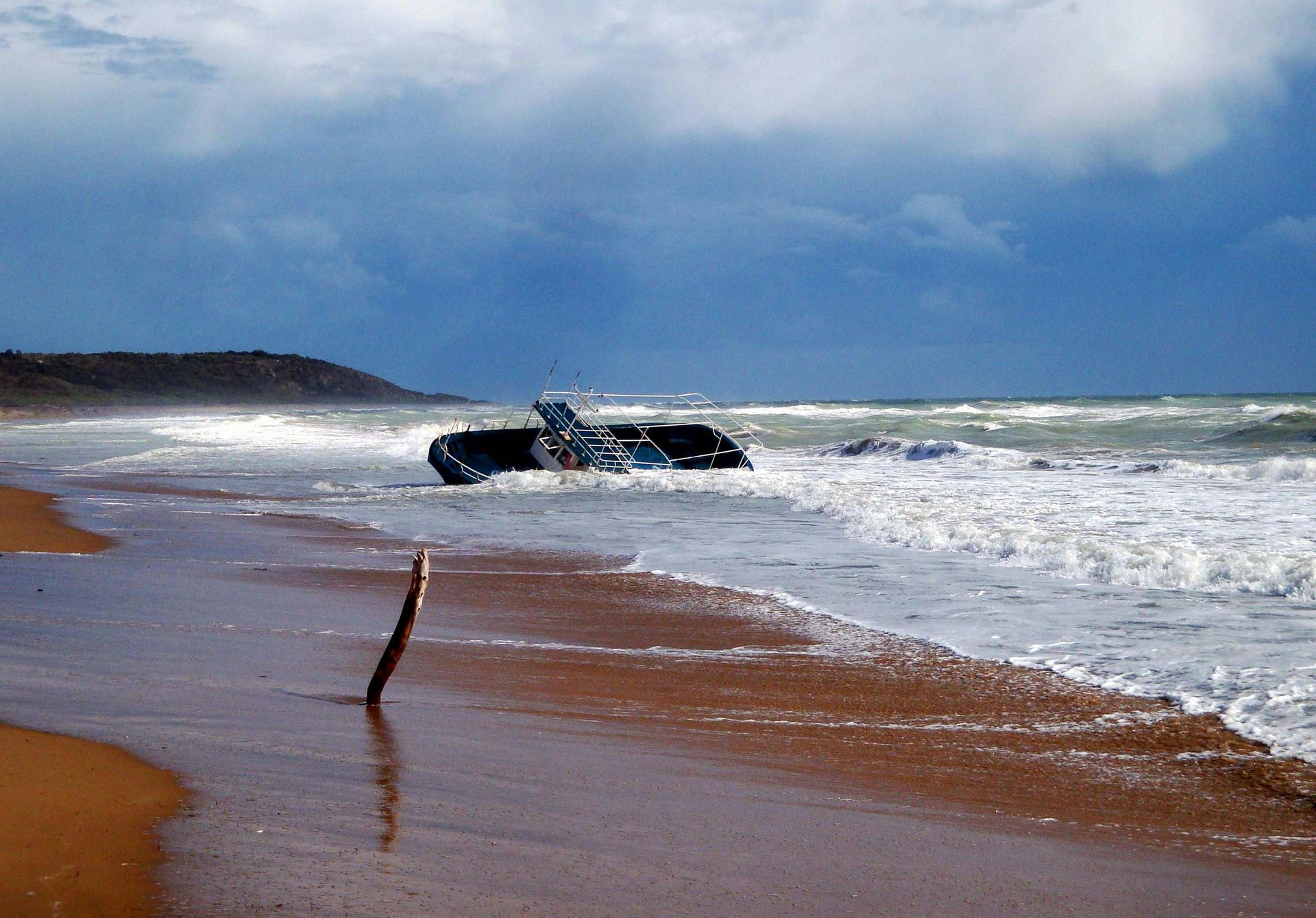 A small boat has washed ashore on a sandy beach under a dark, stormy sky, with waves breaking around it.