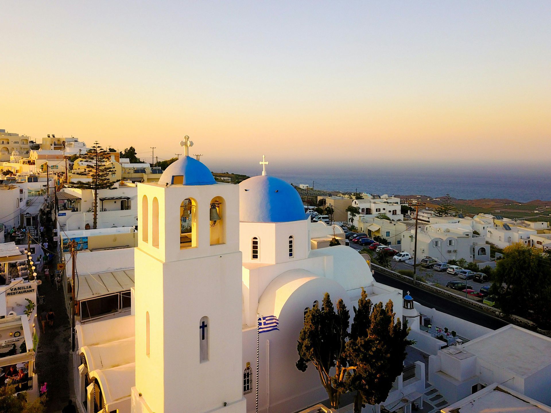 White church with blue domes, Greek flag, and bell tower overlooking a coastal town at sunset.