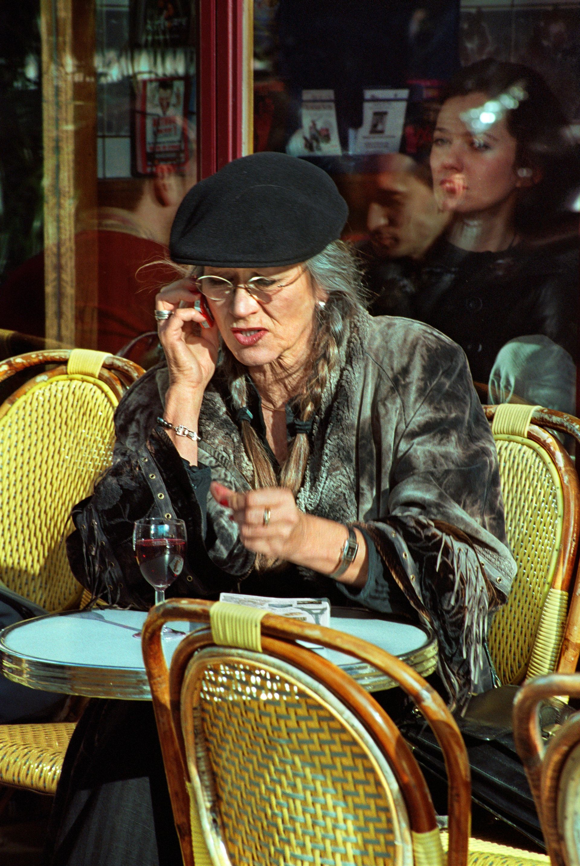 Woman in beret and shawl sits at cafe table, listening. Le Marais