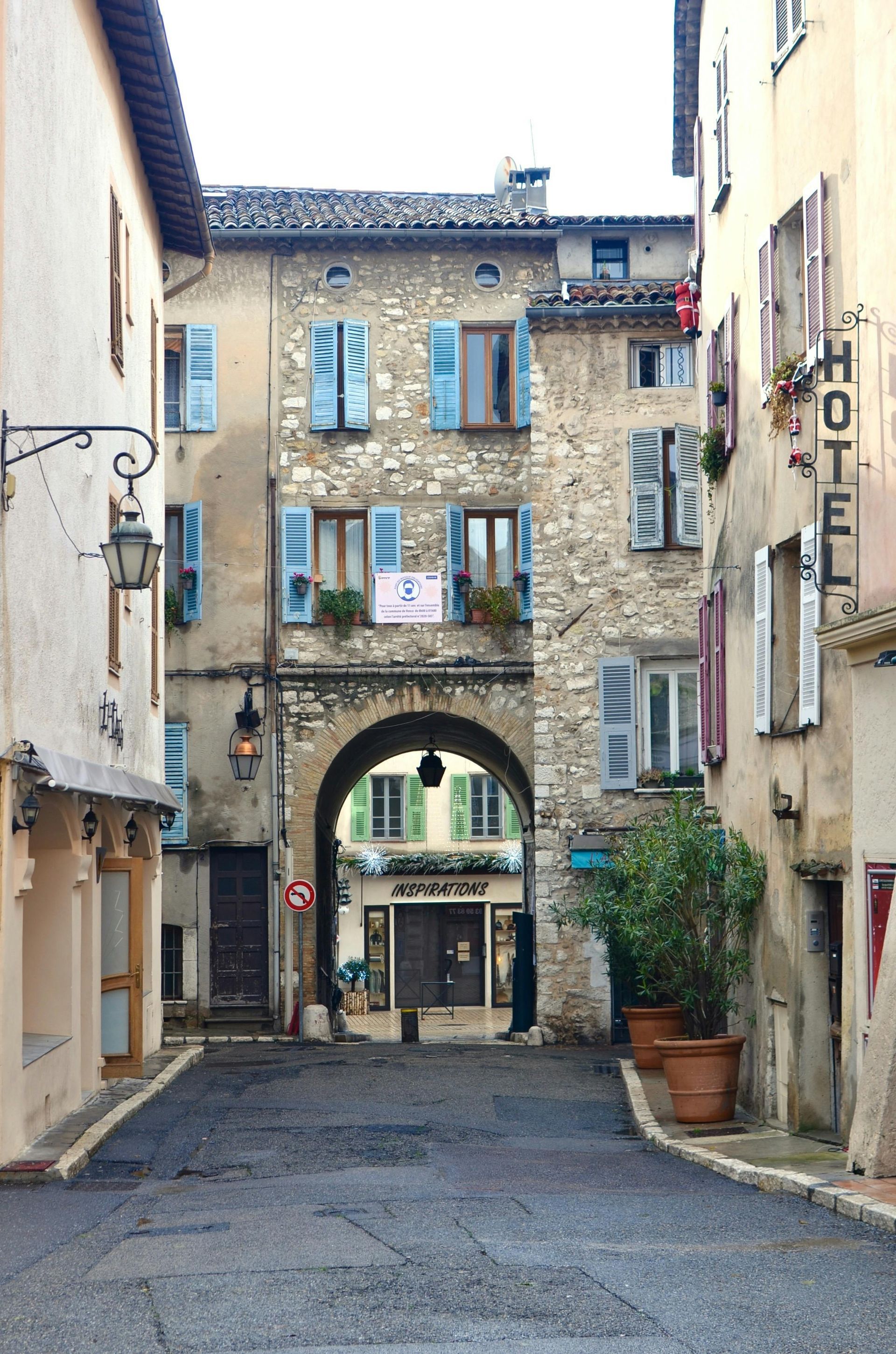 Narrow cobblestone street in a European town with stone buildings, arched entry, and blue shutters.