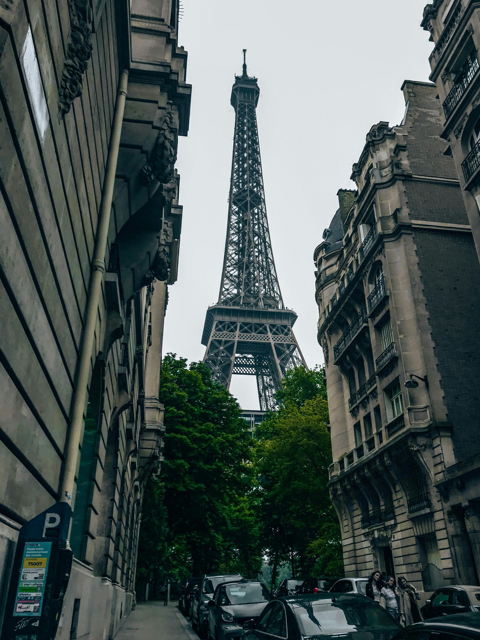 A low-angle view of the Eiffel Tower centered between two tall, ornate Parisian stone buildings on a cloudy day.